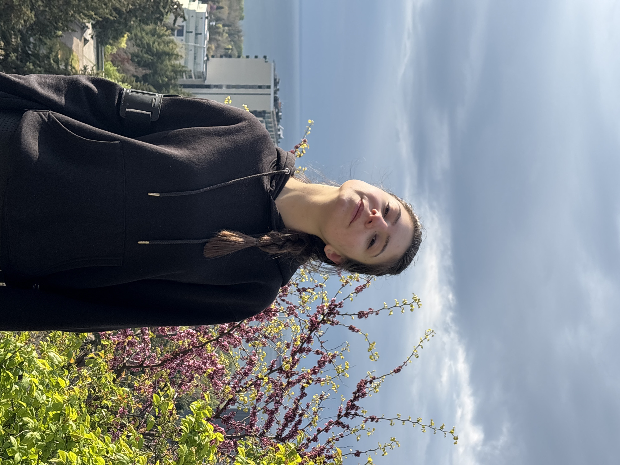 The image shows a young woman with a braided hairstyle, wearing a black hooded sweatshirt. She is standing near a blossoming tree with pink and green leaves. In the background, there are buildings, a body of water, and a cloudy sky. The location appears to be a park or a green space in a city.