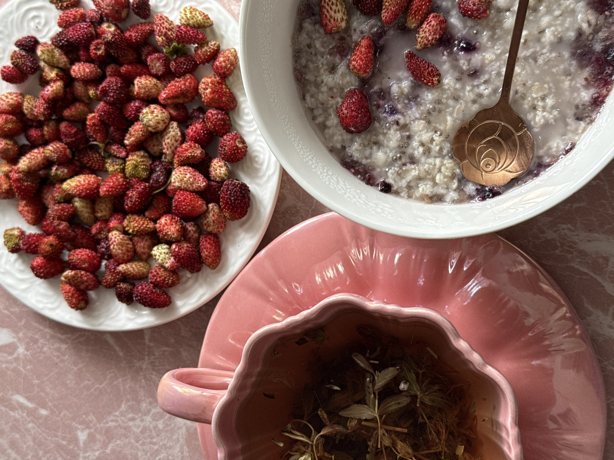 A overhead shot shows a pink marble tablecloth with three dishes of food. On the right, a white bowl contains oatmeal topped with blueberries and small red strawberries. A rose-shaped copper spoon is in the bowl. Above and to the left of the oatmeal bowl is a white plate overflowing with tiny wild strawberries. To the left of the plate of strawberries is a pink scallop-edged bowl filled with dried tea leaves and hot tea.