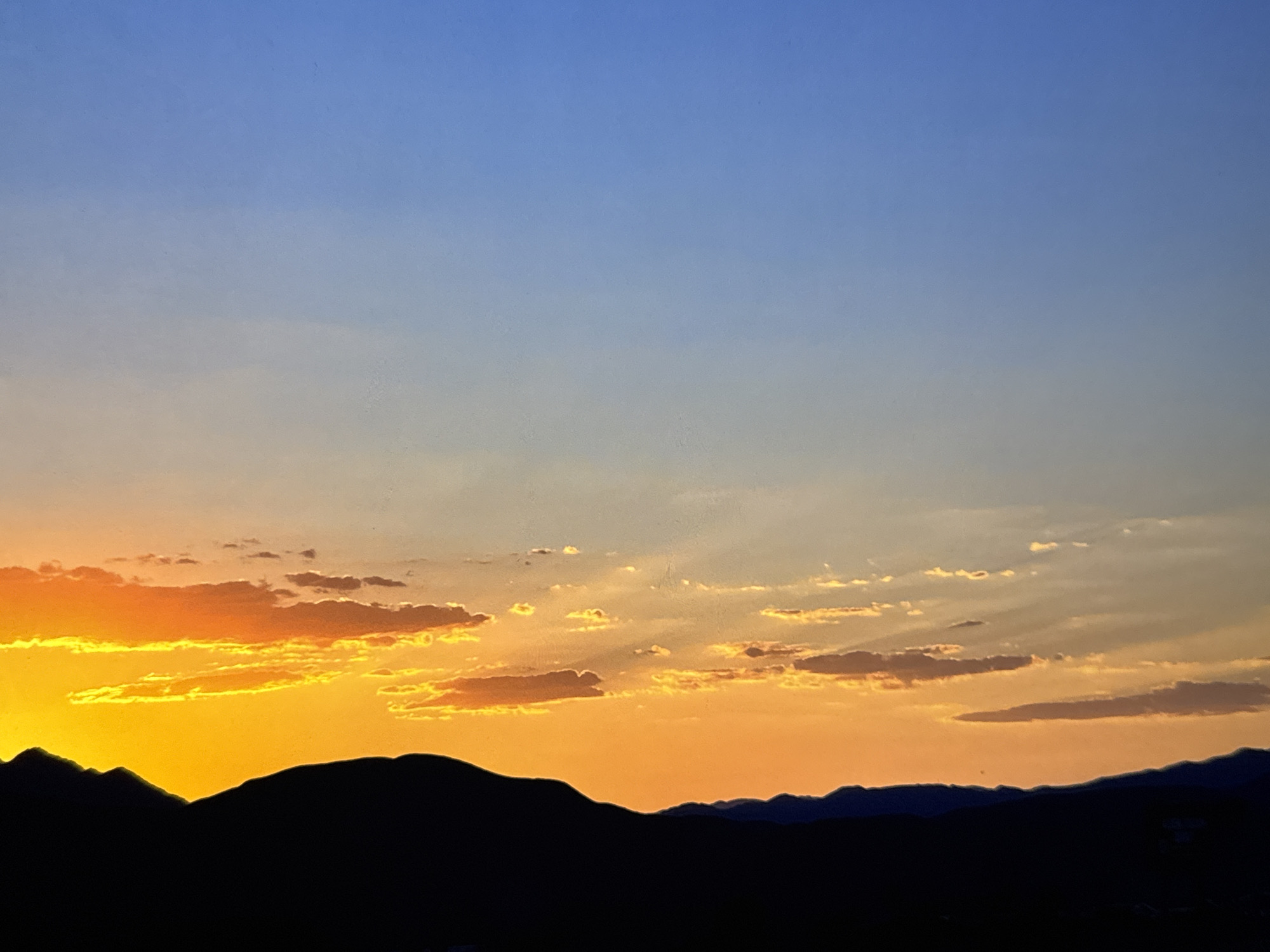 A silhouette of mountain ranges is visible against a vibrant sunset sky. The sky transitions from a deep blue at the top to a bright orange and yellow near the horizon, with scattered clouds illuminated by the setting sun. The image appears to be a landscape photograph taken during twilight. There are no people, animals, or man-made structures clearly visible in the scene, making it difficult to pinpoint an exact location within Los Angeles, United States of America. The environment suggests an outdoor, natural setting.