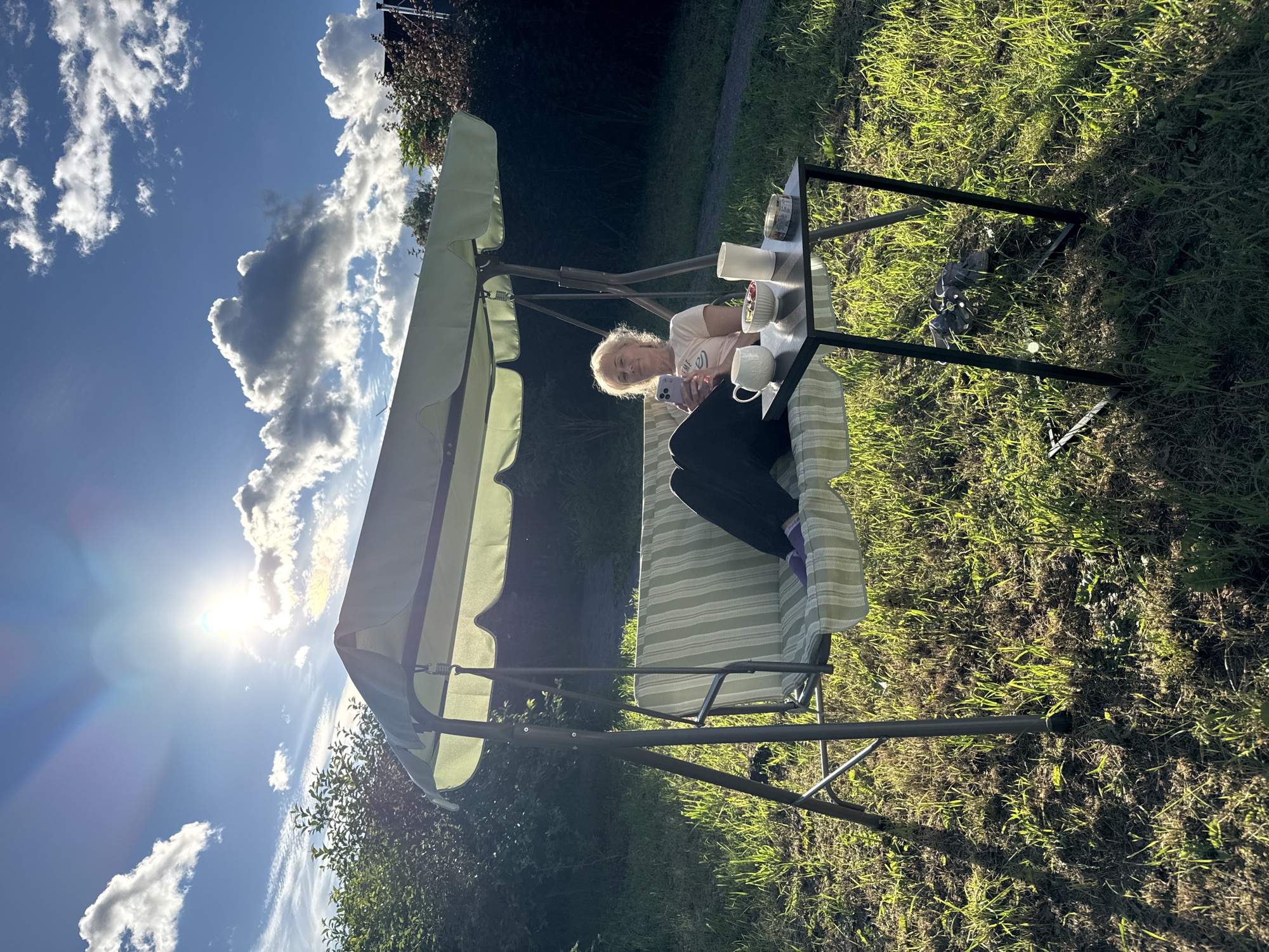 A woman sits on a green and white striped swing, looking at her phone. She wears a light pink t-shirt and dark pants. A small, dark-colored table is next to the swing, with a white mug, a white bowl with red berries, and a white cylindrical object on it. The scene is outdoors in a grassy area, with trees and bushes in the background. The sun is bright and low in the sky, casting long shadows and creating lens flare. The sky is blue with dramatic white and gray clouds. The overall atmosphere suggests a relaxed afternoon or early evening in a backyard or park setting. The image is oriented vertically.