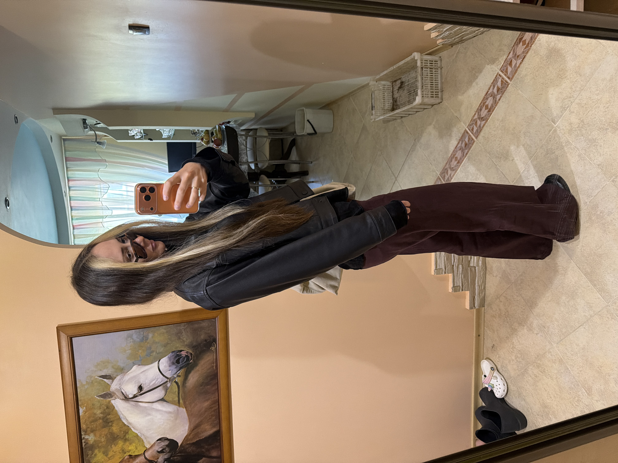 A person stands in a room and takes a mirror selfie. They are wearing a black leather jacket and dark trousers. The room has beige walls, and a framed painting of a white horse hangs on the wall behind the person. The floor is tiled in a diamond pattern. A white plastic basket sits on the floor in the upper-right corner of the image. A staircase is visible on the right side. A white Croc-style shoe lies on the floor. The location is Kamyshin, Russia.
