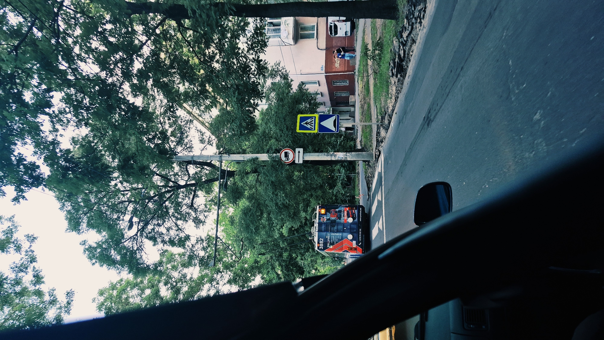 The image captures a street scene in Chișinău, Moldova, looking forward from inside a vehicle. A blue and red trolleybus with the number 3882 prominently displayed is in the left lane, moving away from the viewer. The trolleybus has various advertisements on its side. Ahead of the trolleybus, traffic signs are visible, including a pedestrian crossing sign and a sign indicating a route for vehicles with trailers. A light pole stands tall next to these signs. The street is paved with asphalt and has white markings for a pedestrian crossing. To the right, a multi-story building with light pink walls and white window frames is situated along the street. Several trees with lush green foliage line the road and surround the building, creating a shaded environment. In the foreground, the interior of the vehicle, including a black dashboard and side mirror, is partially visible. A few people are seen walking on the sidewalk near the building. The sky is overcast, and the overall lighting suggests it is daytime.