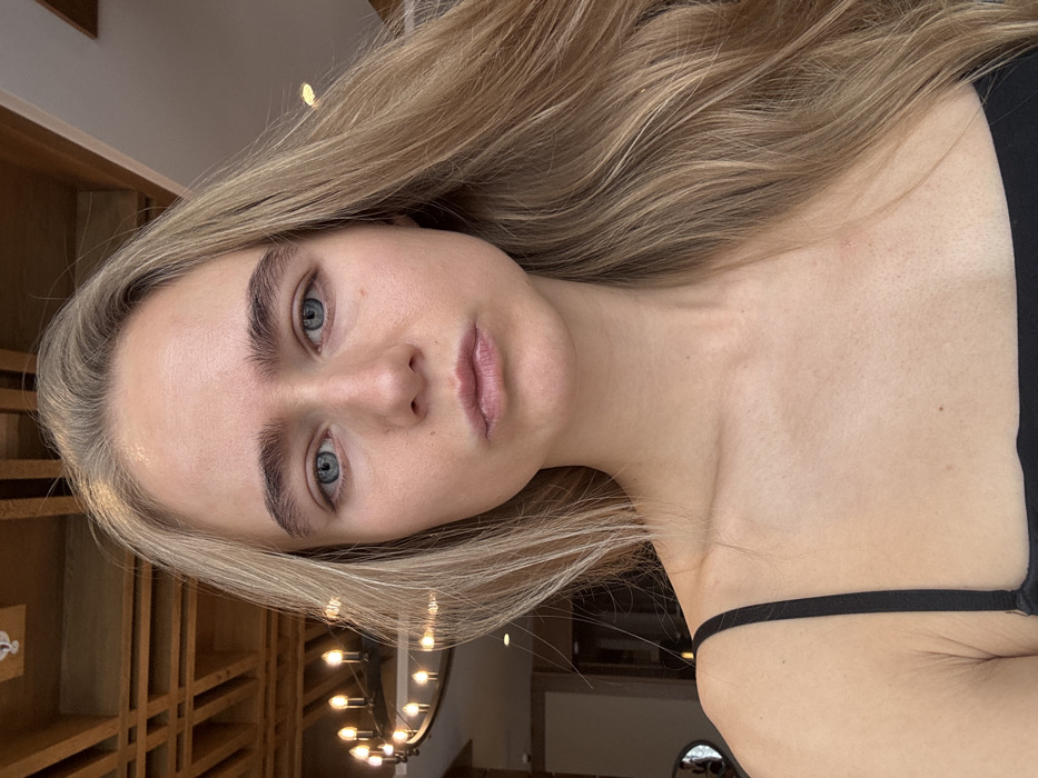 A woman with fair skin, blonde hair, and light blue eyes poses in a selfie-style portrait. She has full eyebrows and lips. A thin black strap is visible over her shoulder. The background shows a wooden wall with decorative molding and a glimpse of a chandelier with exposed light bulbs. The image is rotated slightly counterclockwise.
