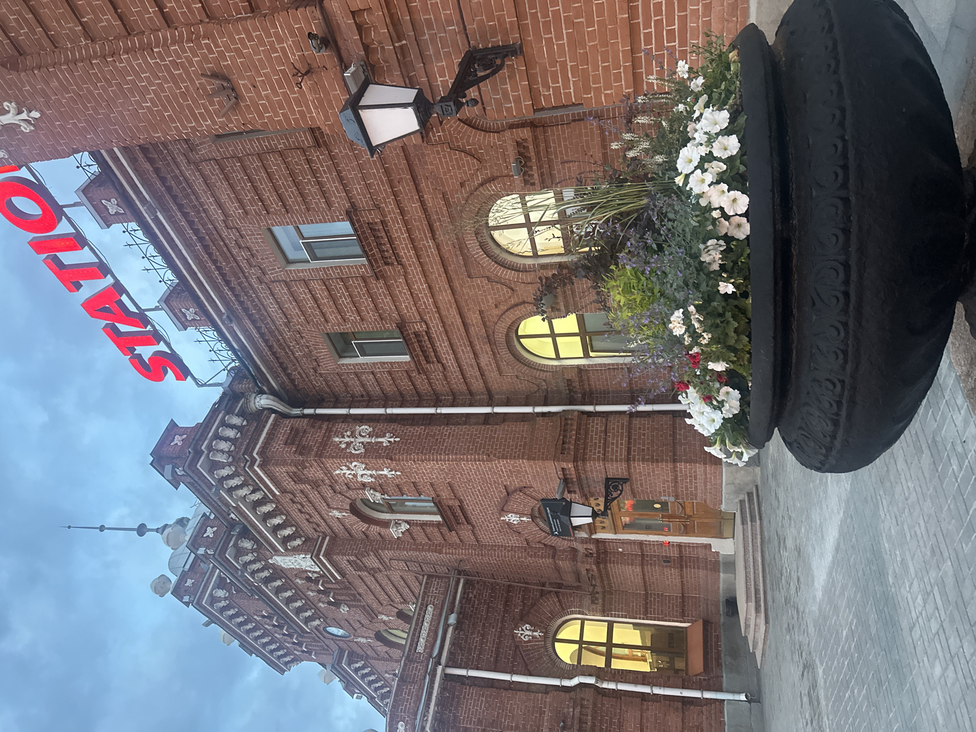 This image depicts the Kazan-Passazhirskaya (Kazan-1) railway station, located in the city of Kazan, Republic of Tatarstan, Russia. The scene captures the building's facade under an overcast, possibly twilight or early morning, sky.
The central subject is a multi-story, ornate building constructed primarily of red brick with extensive white architectural detailing. These white elements include pilasters, cornices, arched window frames, intricate carved motifs, and finials adorning the roofline. A prominent red neon sign spelling "STATION" is mounted on the roof, partially visible at the top center-right. The building features numerous arched windows, some of which are illuminated from within, casting a warm yellow light. A small, circular clock face is visible on the upper left section of the facade. A tall, slender spire with an antenna-like top protrudes from the central roof structure. White downspouts run vertically along the brick walls.
In the foreground, on the right side, there is a large, dark-colored, intricately carved planter filled with various plants, including white petunias, purple flowering plants, and green foliage. An ornate, black, wall-mounted lamp with a white diffuser is affixed to the brick wall on the right. The ground immediately in front of the building is paved with light grey interlocking pavers. A set of three steps leads up to a recessed entrance featuring double wooden doors. No people or animals are visible in the image.