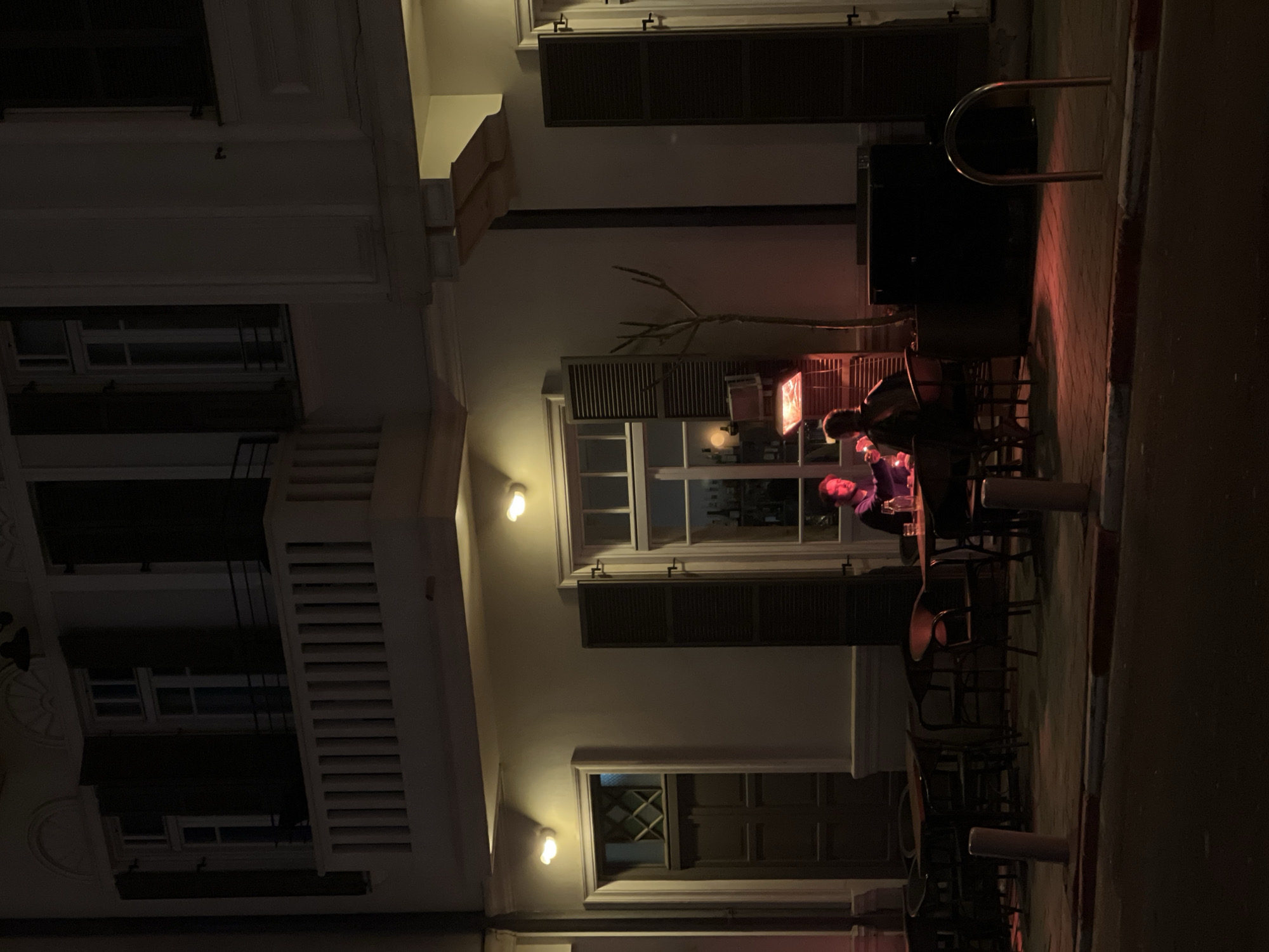 The image depicts a nighttime scene on a street in Tel Aviv, Israel. A two-story building with a light-colored facade occupies the background, featuring multiple windows with dark louvered shutters. A white balustraded balcony is present on the second floor. At street level, an outdoor dining area is established on a paved sidewalk.
Two individuals are seated opposite each other at a small, dark table. The person on the left has reddish hair and is seen in profile, illuminated by a red glow. The person on the right has dark hair and is partially obscured. A red-glowing patio heater is positioned adjacent to their table, casting a prominent red light on the area. Bottles and glasses are visible on the table surface.
Wall-mounted sconces on the building illuminate portions of the facade with a warm yellow light. Through the windows, a bar area with bottles on shelves is discernible. To the left of the dining area, additional dark tables and chairs are arranged. A small, bare tree or shrub stands in a planter near the illuminated table.
The foreground consists of a section of an asphalt street, bordered by a curb painted with red and white stripes. Two light-colored cylindrical bollards are embedded in the sidewalk, and a silver U-shaped bike rack is present towards the right side of the frame.