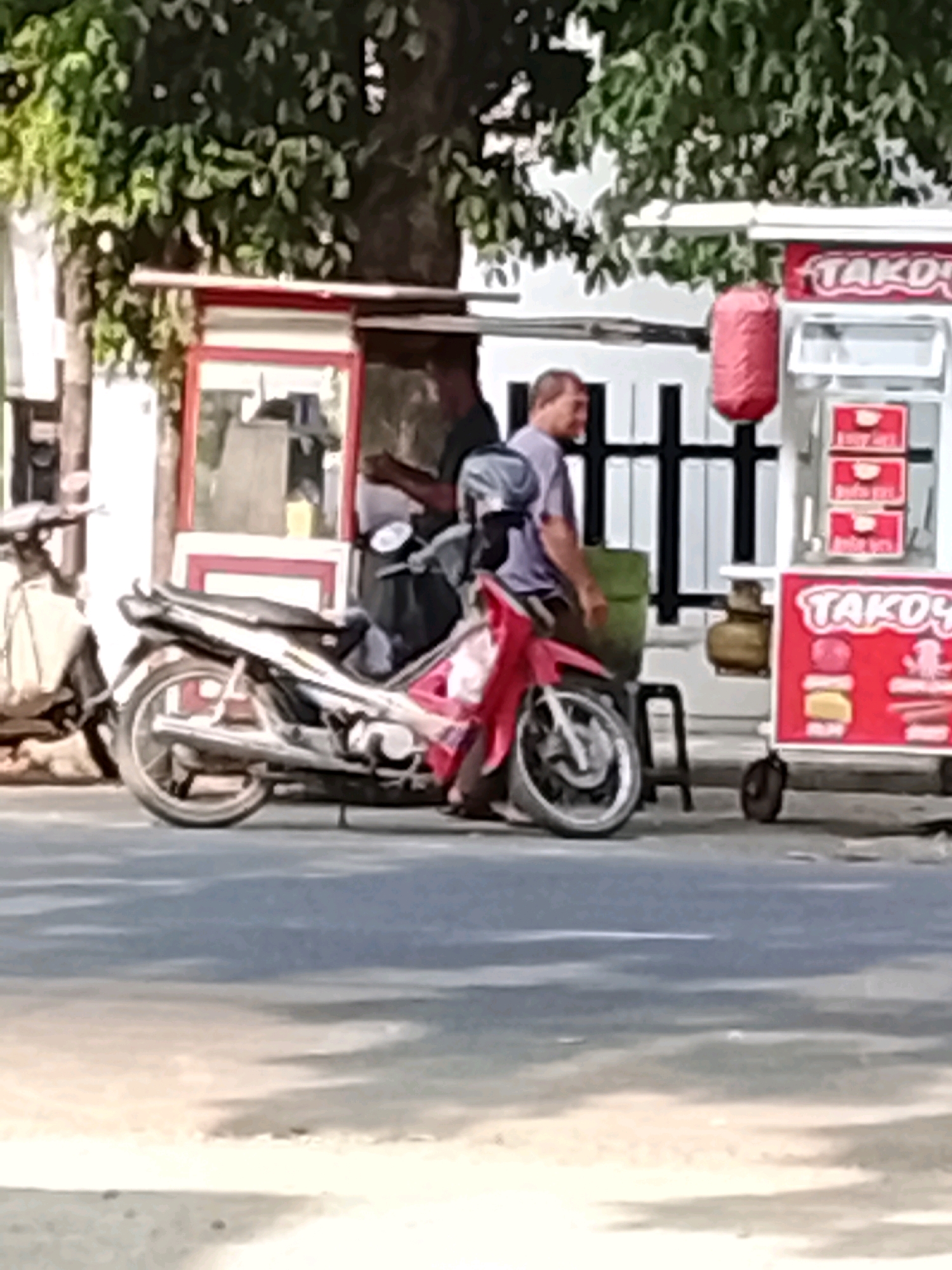The image depicts a street scene in Slawi, Indonesia, likely near a food stall. In the foreground, a red and black motorcycle is parked. Behind the motorcycle, a man in a light purple shirt stands next to a green container and a food cart. The food cart, identified by the sign "TAKOS," has a red and white exterior and displays red boxes with white lettering, possibly indicating menu items. Another food cart is partially visible to the left, with a red and white frame and a glass display. The background shows a tree with green foliage and a white structure with black bars. The ground is paved with asphalt. The lighting suggests it is daytime.