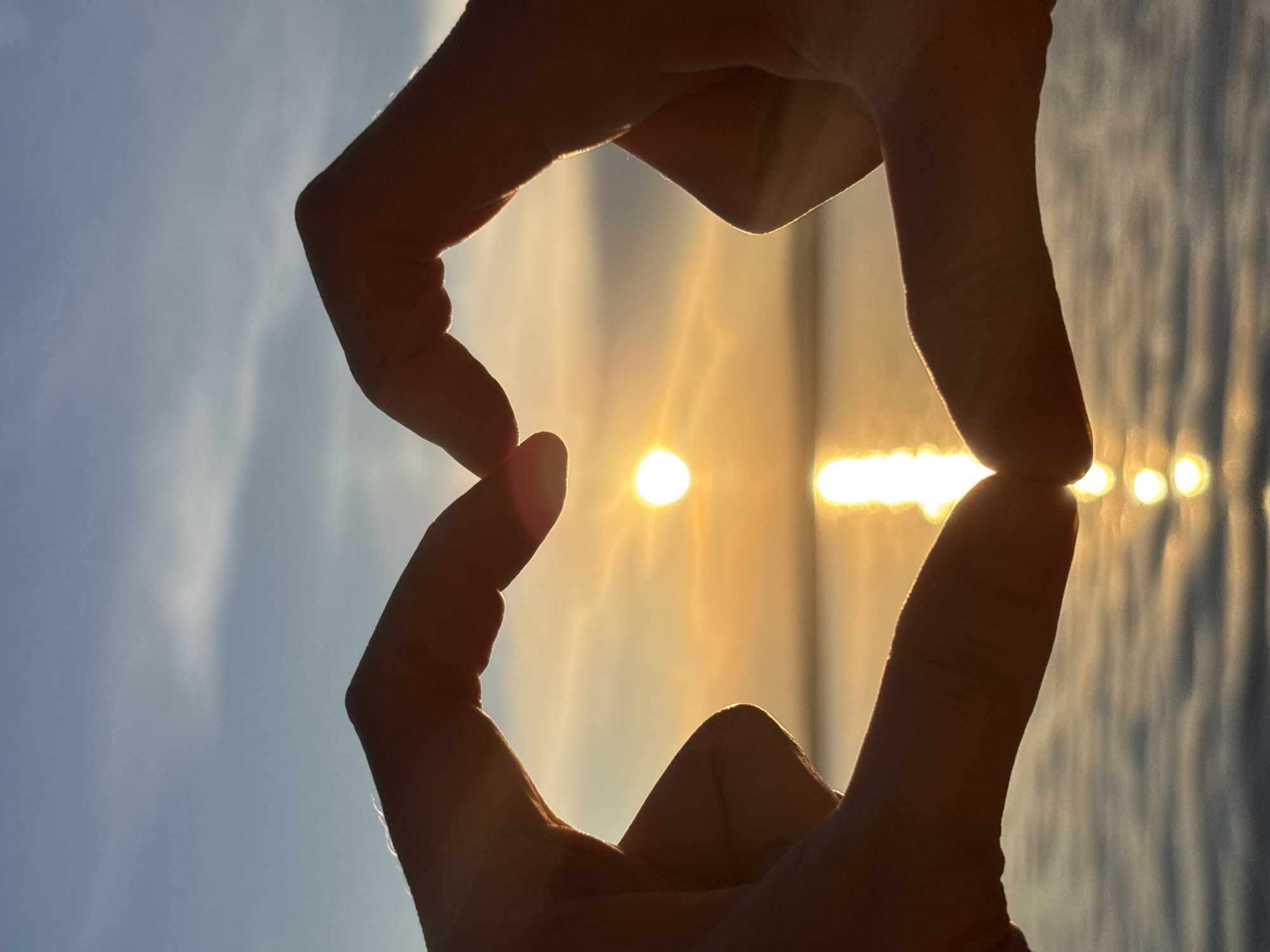The image displays a close-up silhouette of two hands forming a heart shape against a sunset over a body of water. The sun is partially visible, appearing as a bright, warm orb. The sky is a gradient of blues and oranges, with soft clouds. The water reflects the sunset with shimmering streaks of light. The location is identified as Unknown, Unknown. The focus is on the hands creating the heart shape, with the sunset and water serving as the background.