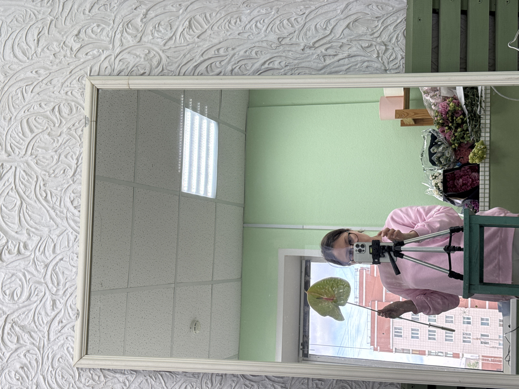 An indoor mirror selfie is captured, with the image rotated 90 degrees clockwise, causing the reflection of the person and interior elements to appear sideways. The mirror has a white, decorative frame and is mounted on a wall covered with textured white wallpaper featuring swirling patterns.
Reflected in the mirror, a person with dark hair, glasses, and a clear face shield is seen wearing a pink long-sleeved top. They are holding a smartphone mounted on a small tripod with their left hand and a large, green-red anthurium leaf or flower on a long stem with their right hand. The person appears to be posing for a photo or video.
The reflected background reveals a light green wall and a white drop ceiling with fluorescent lighting. To the right of the reflected person, a section of the green wall features wooden shelves holding rolls of pink and white paper, alongside various bouquets of flowers wrapped in plastic, suggesting a flower shop or studio environment. Through a window, a multi-story building with red/brown and light-colored facades and numerous windows is visible under a cloudy daytime sky. The setting is in Izhevsk, Russia.