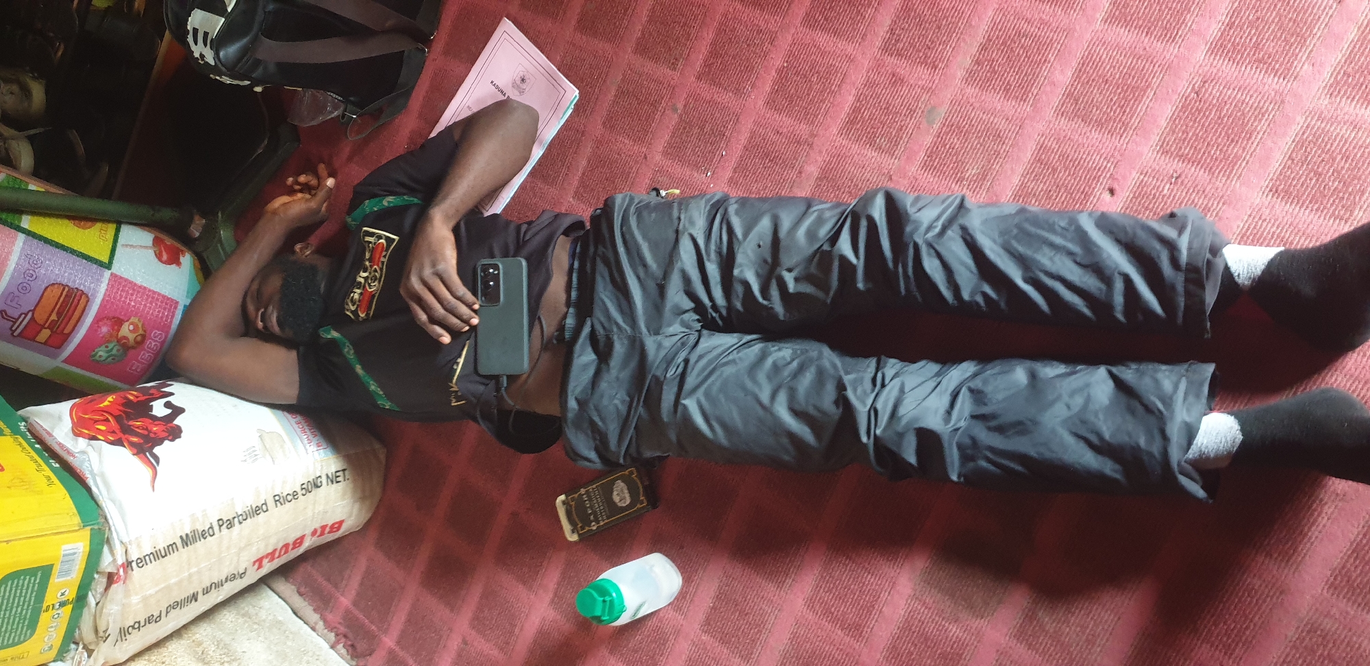 A Black male is lying on his back on a red carpeted floor. He is wearing a black t-shirt with green trim and gold accents, and black pants. A smartphone is on his chest. Next to him are a plastic bottle with a green lid and a small rectangular object. In the background, there are large bags of rice and other items. The scene appears to be indoors, possibly in a residential or storage area in Sabon Tasha, Nigeria.