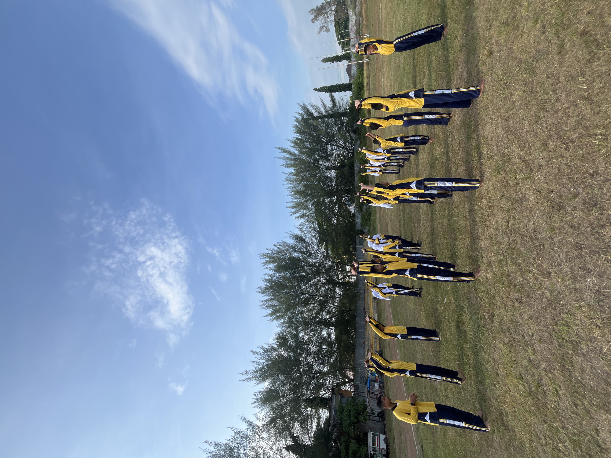 A group of approximately 20 students, dressed in matching yellow and dark blue athletic uniforms with white accents, are performing synchronized exercises on a grassy field. They are arranged in multiple rows, facing away from the camera, with their arms raised and extended overhead. The field appears to be part of a sports complex, with a track and stadium seating visible in the background. Behind the field are rows of tall, slender trees, and beyond that, buildings and a white van. The sky above is a clear blue with scattered white clouds. This scene is likely taking place in Lhokseumawe, Indonesia, given the presence of typical Indonesian flora and architecture.