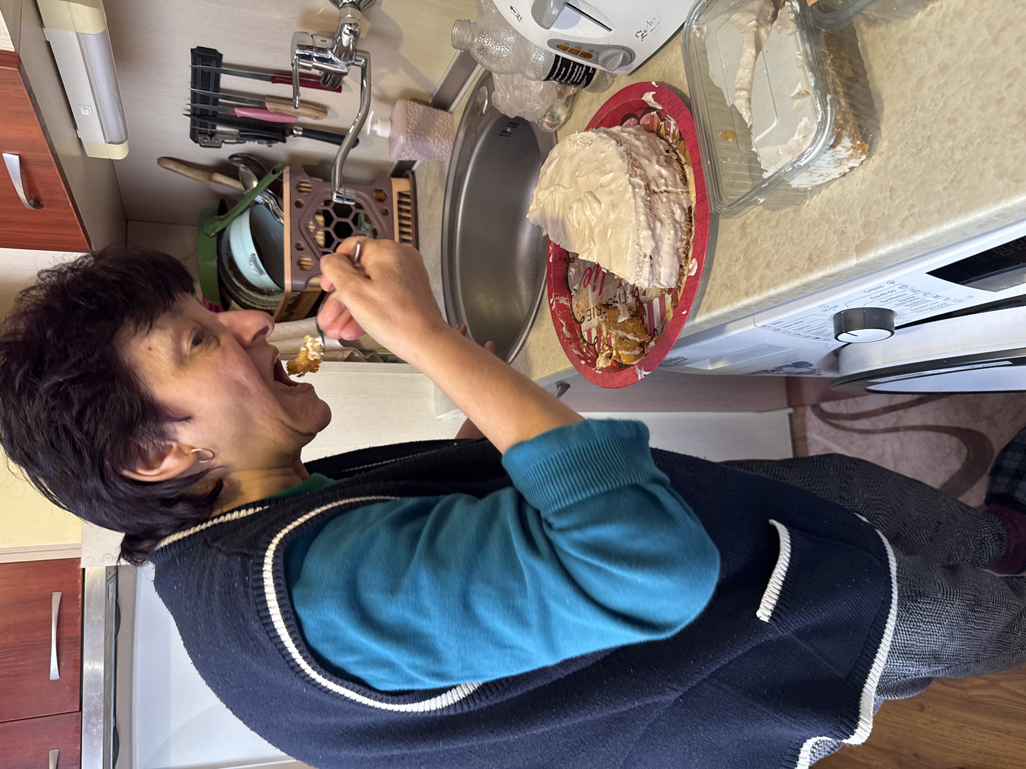 A person with dark hair is eating cake from a fork while standing in a kitchen. The kitchen features wood cabinets with silver handles and a stainless-steel sink. There's a drying rack above the sink and a white microwave to the right of the sink. The person is wearing a teal sweater and a dark blue vest with white trim. Additional cake is in a clear plastic container. The image appears to have been taken indoors. The location is not verifiable as Burgas, Bulgaria, based on the image.