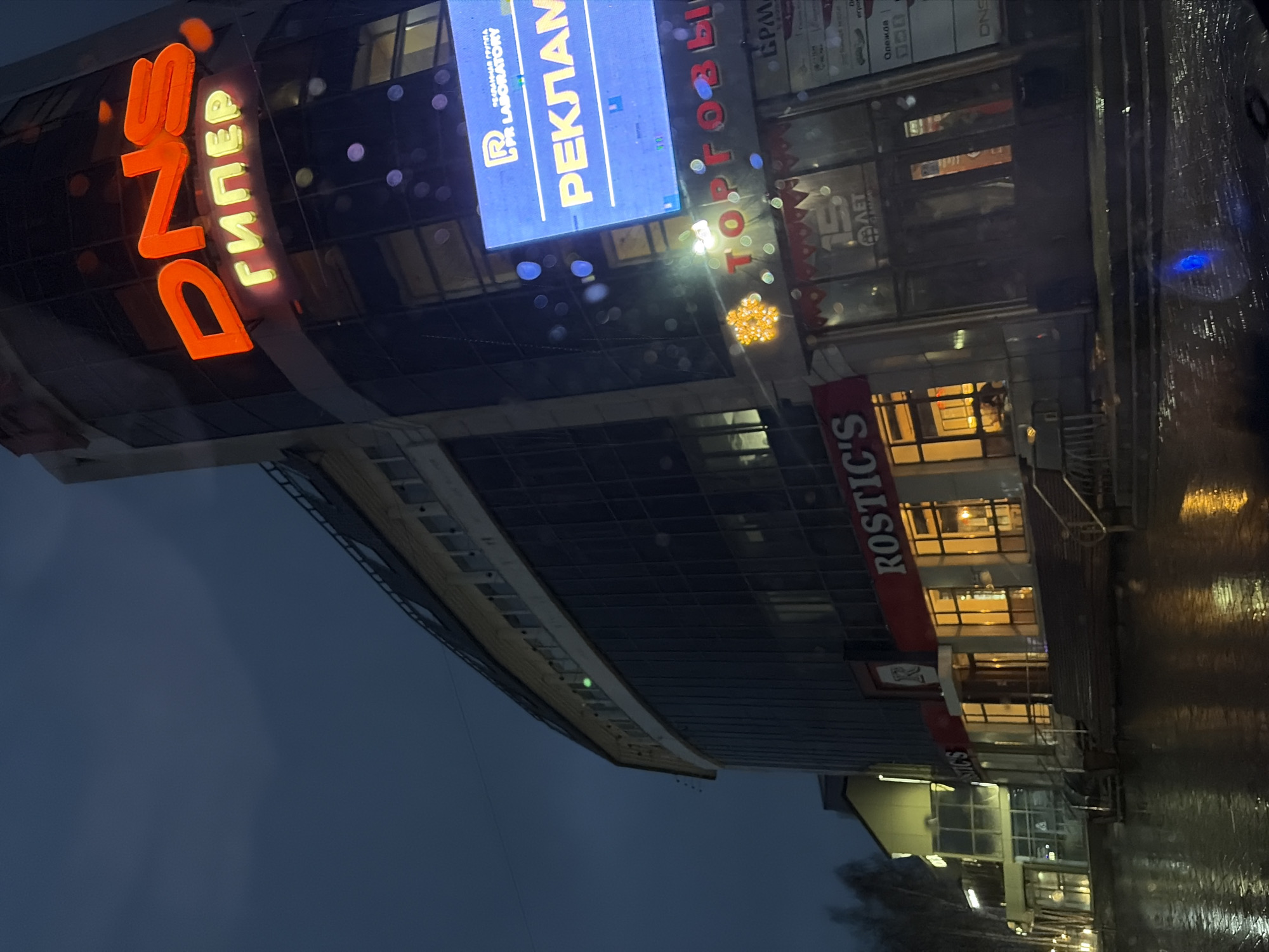 The image captures an illuminated commercial building in Chaikovsky, Russia, at dusk or night, under conditions suggesting recent rain. The central focus is a modern, multi-story structure featuring a curved glass facade.
Prominently displayed on the building's left side is a large orange and yellow neon sign spelling "DNS ГИПЕР" (DNS HYPER). Above this, a large blue digital display screen shows text, including "РЕКЛАМНАЯ ГРУППА PR LABORATORY" (ADVERTISING GROUP PR LABORATORY) and the partial word "РЕКЛАМ" (ADVERTIS). Further right, a red banner sign reads "ROSTIC'S". Other visible Cyrillic text includes "ТОРГОВЫЙ" (TRADING / RETAIL), "15 ЛЕТ" (15 YEARS), "GPM," "Олежав," and "игров."
The ground or street in front of the building is wet, reflecting the vibrant artificial lights from the various signs and interior windows, creating a shimmering effect. A decorative, star-shaped light fixture is also visible on the building's exterior. No people are visible in the frame. The scene conveys an active urban commercial area illuminated against a dark evening sky, with the wet surfaces adding to the atmospheric conditions.