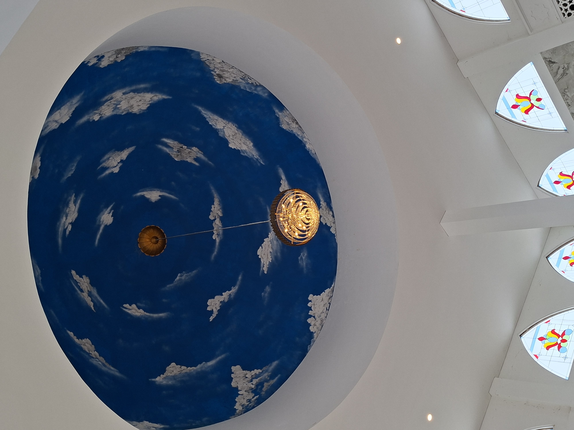 The image captures the interior of a building in Keureuweungkrueng, Indonesia, focusing on a prominent dome-shaped ceiling. The dome is painted with a dark blue background and scattered, stylized white cloud formations, creating a sky-like effect. Two light fixtures hang from the center of the dome, one a smaller, simple structure and the other a more elaborate, multi-tiered chandelier. To the upper right of the image, sections of the ceiling are visible with decorative stained glass windows in a semi-circular shape, featuring red, yellow, and blue floral designs. The surrounding ceiling is white, with clean architectural lines and curves.
