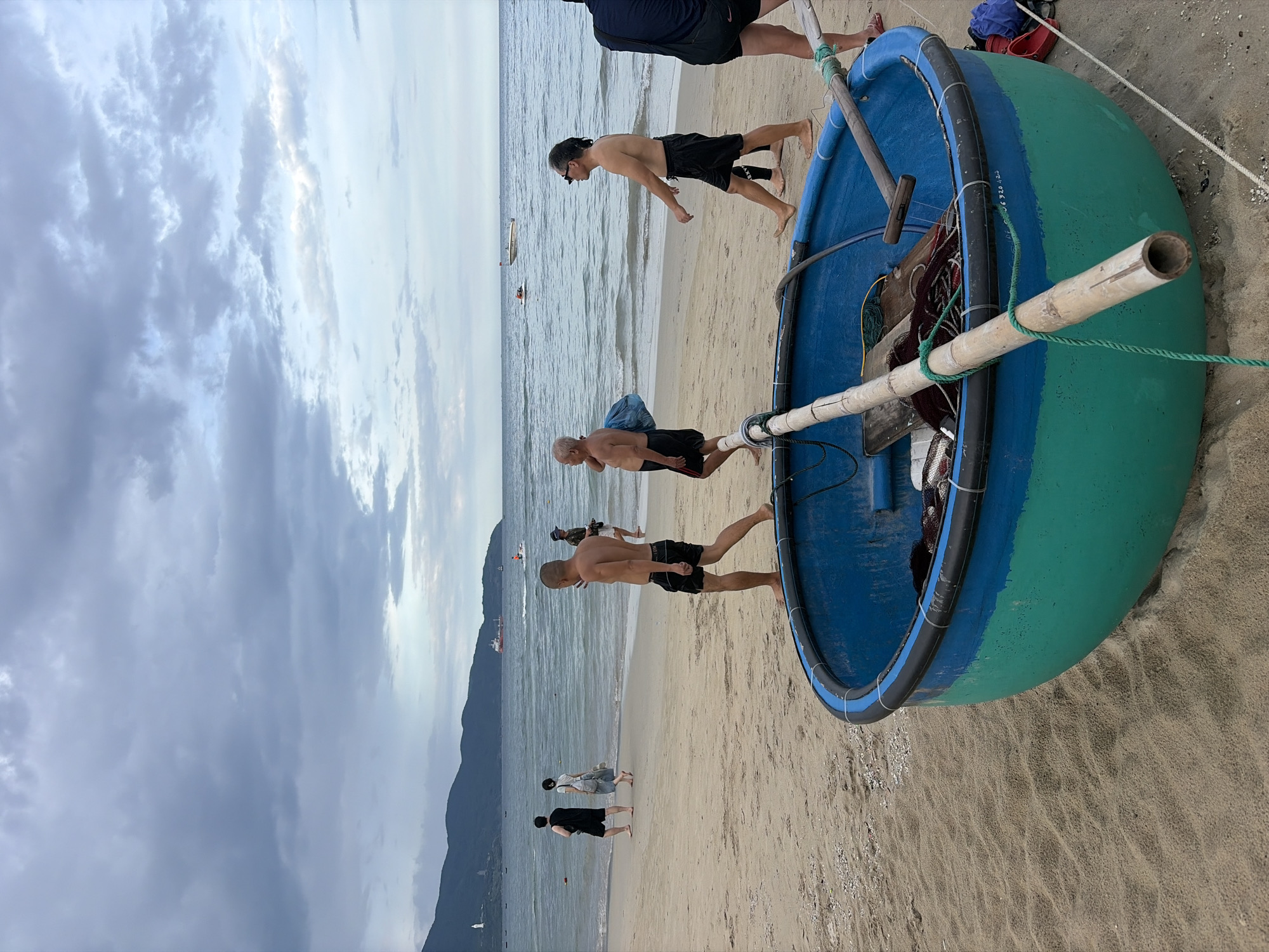 The image depicts a beach scene in Đà Nẵng, Vietnam. In the foreground, a blue and green coracle boat is partially visible, resting on the sandy beach. Several people are walking along the shoreline in the middle ground. In the distance, the ocean stretches to the horizon, with a few boats and mountains visible. The sky is overcast with clouds, suggesting a potentially cloudy day or evening. The overall atmosphere is relaxed, with people enjoying a stroll on the beach.