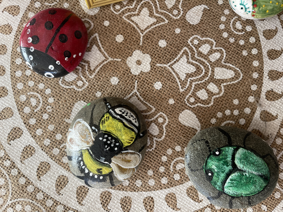 This media file presents a close-up view of three distinct hand-painted rocks, artfully arranged on a light brown, woven fabric featuring white, ornate patterns, suggesting a decorative placemat or tablecloth. The scene is set indoors, likely on a flat surface such as a table.
In the upper left quadrant, a smooth, oval-shaped rock is vibrantly painted as a red ladybug with black spots, a black head, and small white details representing eyes and antennae. Centrally positioned towards the bottom-left, another rock depicts a yellow and black bee, complete with delicate white wings and antennae-like details, set against a grey background. To the right, in the lower corner, the third rock showcases a green insect, possibly a beetle or grub, characterized by its bright green body, black outlines, and subtle white highlights, integrated into a darker grey, textured design that mimics its natural habitat.
A small, light-colored wooden frame is partially visible in the upper center, hinting at other decorative elements nearby. There are no people present, and no active event is taking place; the image captures a static display of these crafted items. The bright, even lighting suggests it is daytime. The location context provided for this scene is Castione, Italy. No visible text appears within the image.