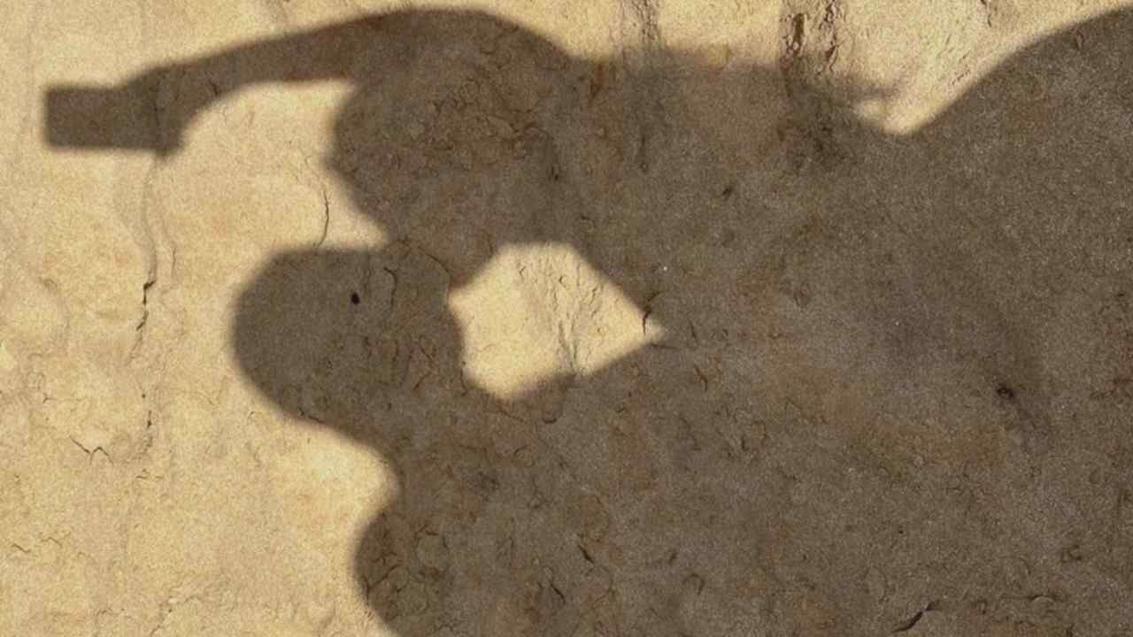 The image displays a shadow of two people kissing, projected onto a sandy, textured surface. One individual, presumably holding a phone, casts a shadow with an arm raised, creating a silhouette that suggests they are taking a selfie. The other figure's shadow is positioned close, with their heads touching in a kissing embrace. The overall impression is a romantic moment captured in silhouette against a natural, earthy backdrop. The location is unknown.