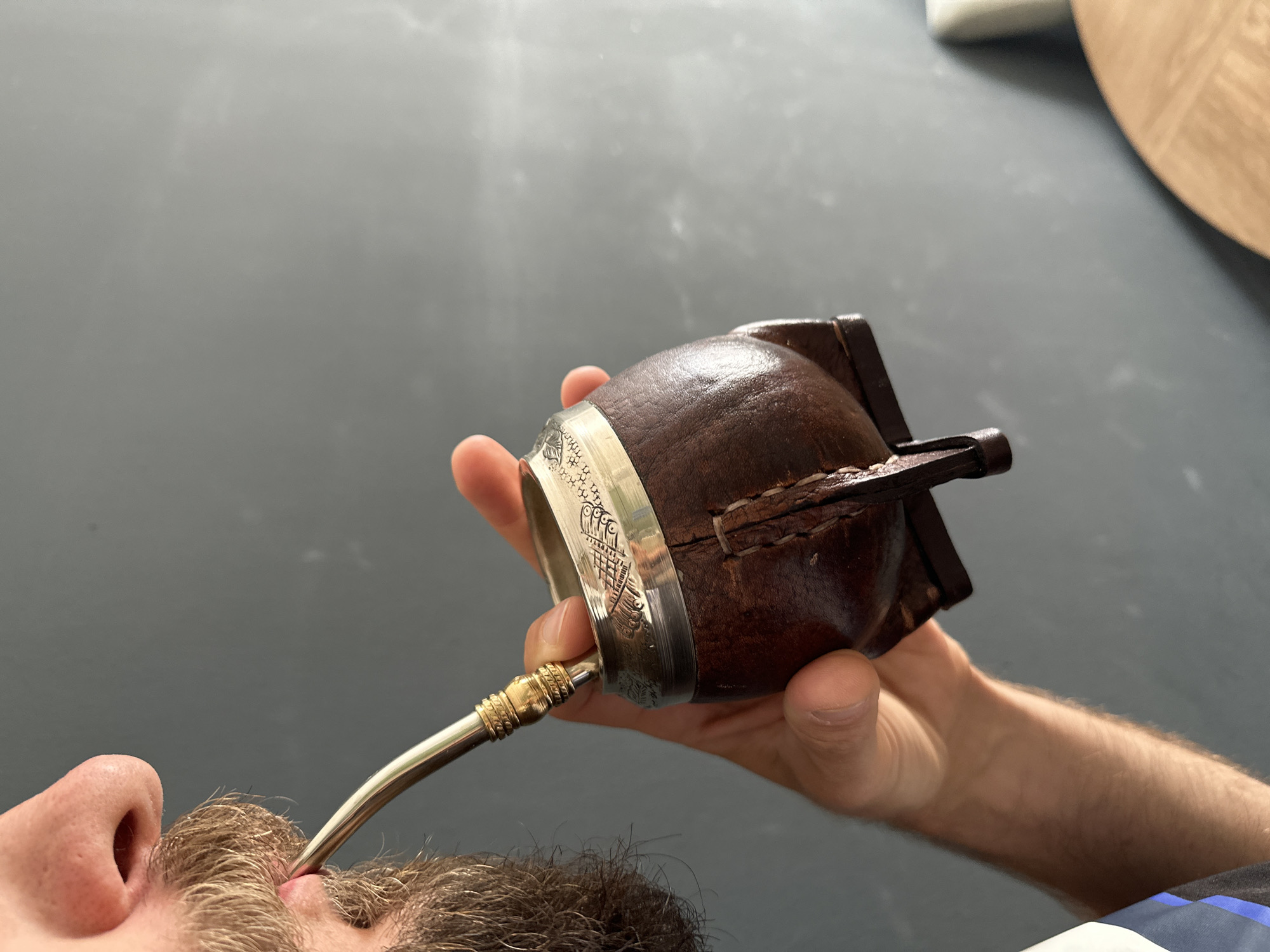 An eye-level, slightly tilted shot captures a person engaged in the act of drinking Yerba Mate. The scene is set indoors, against a dark grey, subtly textured surface that likely serves as a tabletop or counter. In the blurred background to the upper right, parts of a light-colored wooden circular object and a white object are visible.
The primary subjects are a person and the mate drinking set. The lower left portion of the image shows the person's nose, mouth, and a section of their beard or stubble. A light-skinned hand, presumably belonging to the same individual, firmly grasps a dark brown mate gourd. This gourd has a rustic appearance, possibly crafted from wood or calabash and covered with stitched leather, and is adorned with a prominent silver-colored metal rim featuring intricate, engraved patterns. A silver bombilla, or drinking straw, with a distinct golden decorative band near its top, extends from the gourd to the person's mouth.
The activity clearly depicted is the consumption of Yerba Mate, a traditional South American beverage. The lighting in the image is bright and even, indicating a daytime setting. No specific location cues are visible within the frame, but the context provided places the scene in Fethiye, Turkey. There is no visible text in the image, and due to the partial view of the person's face, no specific emotions are discernible beyond the relaxed nature of the activity.