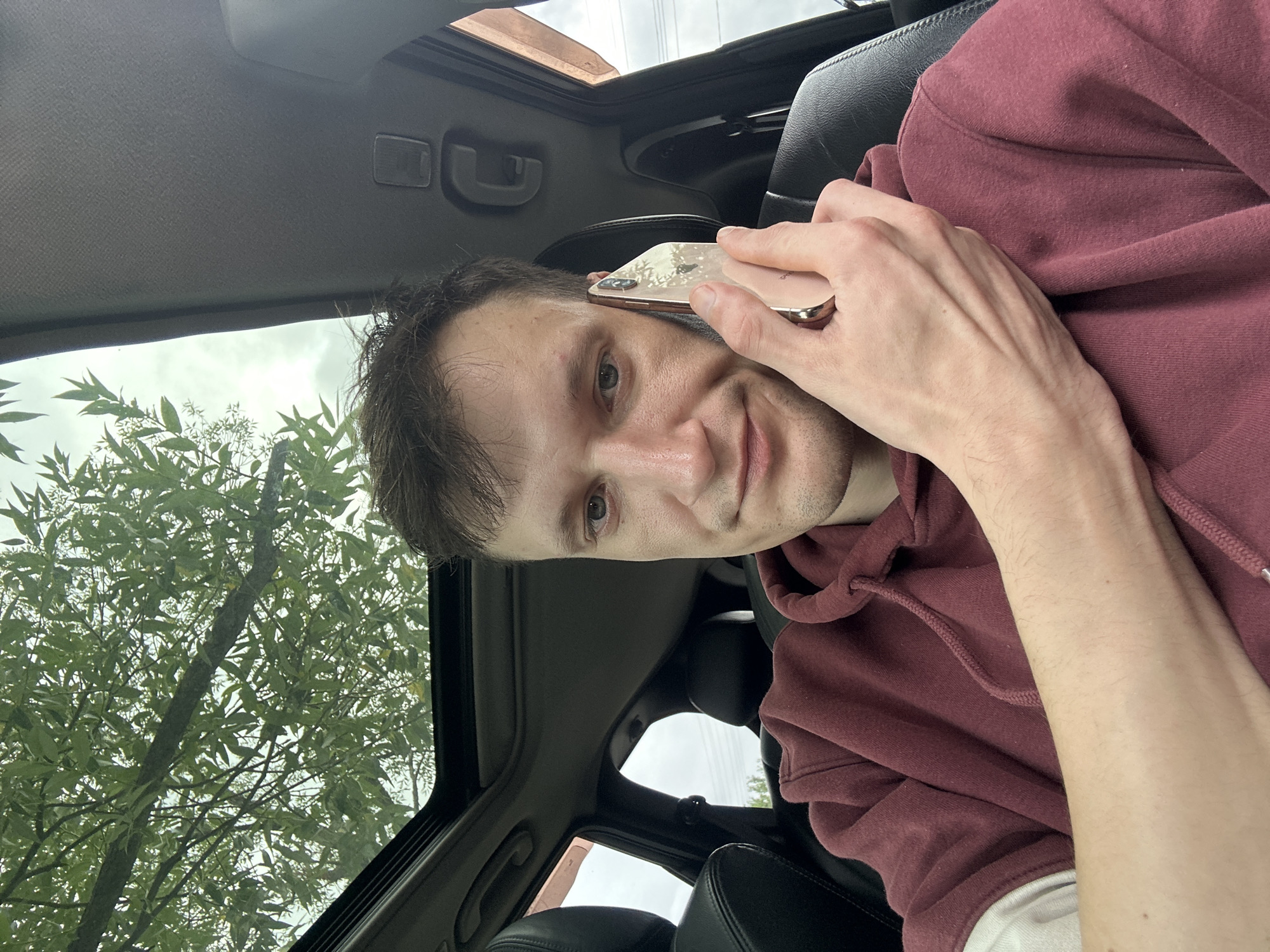 A man with short brown hair, wearing a maroon hoodie, is sitting in the driver's seat of a car, holding a cell phone to his ear with his right hand. The car has a sunroof, through which green tree branches and a cloudy sky are visible. The interior of the car appears to be dark, with black leather seats. The man is looking at the camera with a neutral expression. The image was taken in the city of Makiv, Ukraine.