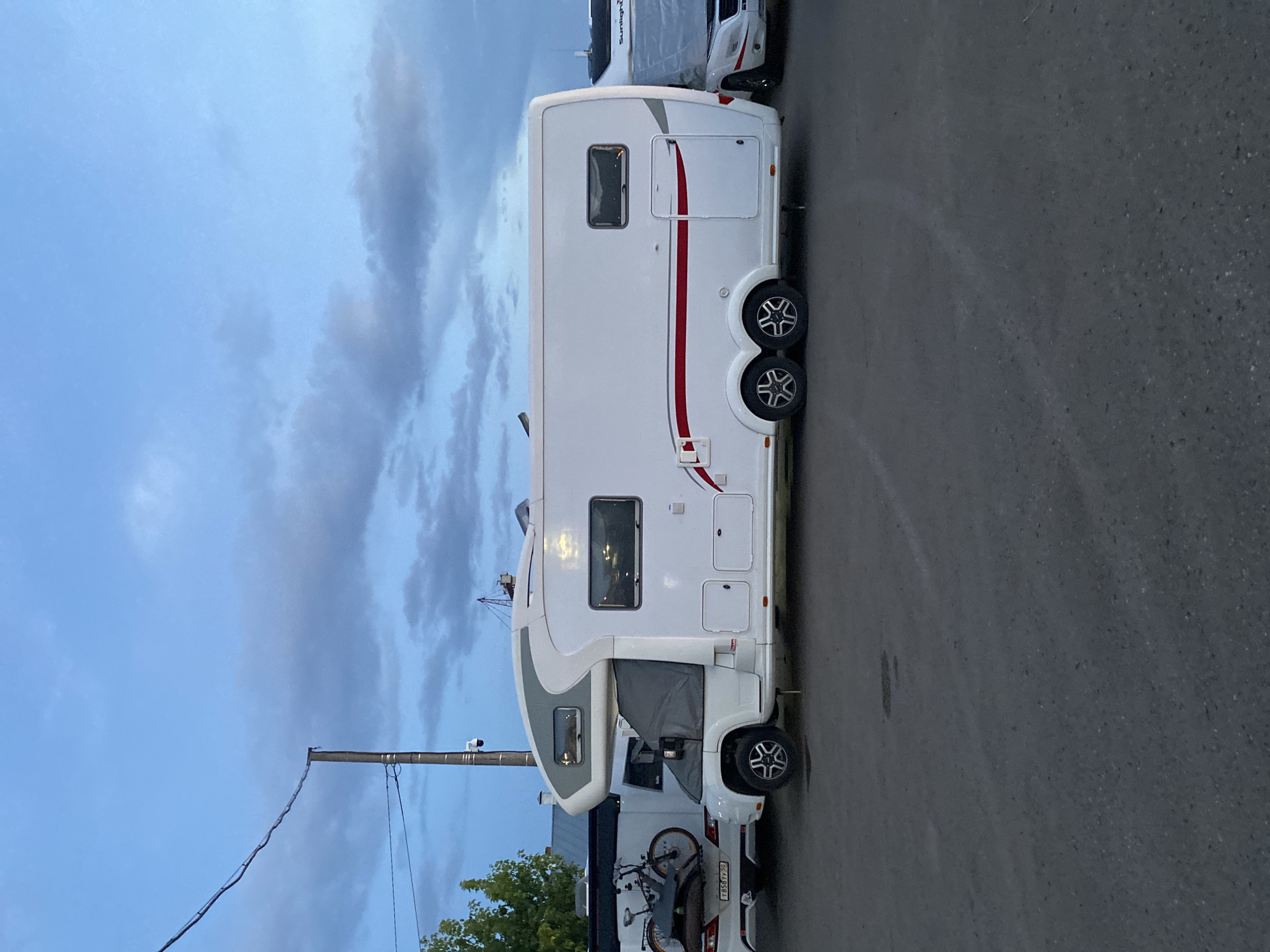 This media file shows a side view of a white motorhome parked on an asphalt surface. The motorhome has a prominent grey stripe along its front section and a red stripe accenting its side. It has multiple windows, including a large one at the front and two smaller ones along the side. There are dual wheels on the rear axle of the motorhome.
Behind the motorhome, another white vehicle is partially visible, with a bicycle mounted on its rear. The license plate of this rear vehicle is visible and appears to read "T 954TY 63".
The setting is outdoors, with a cloudy sky overhead that suggests it is either early morning or late evening. The lighting is dim, consistent with twilight. The environment appears to be an urban or semi-urban area, with a utility pole and wires visible to the left, and some greenery on the far right. No people are visible in the frame. The overall mood is quiet and still.