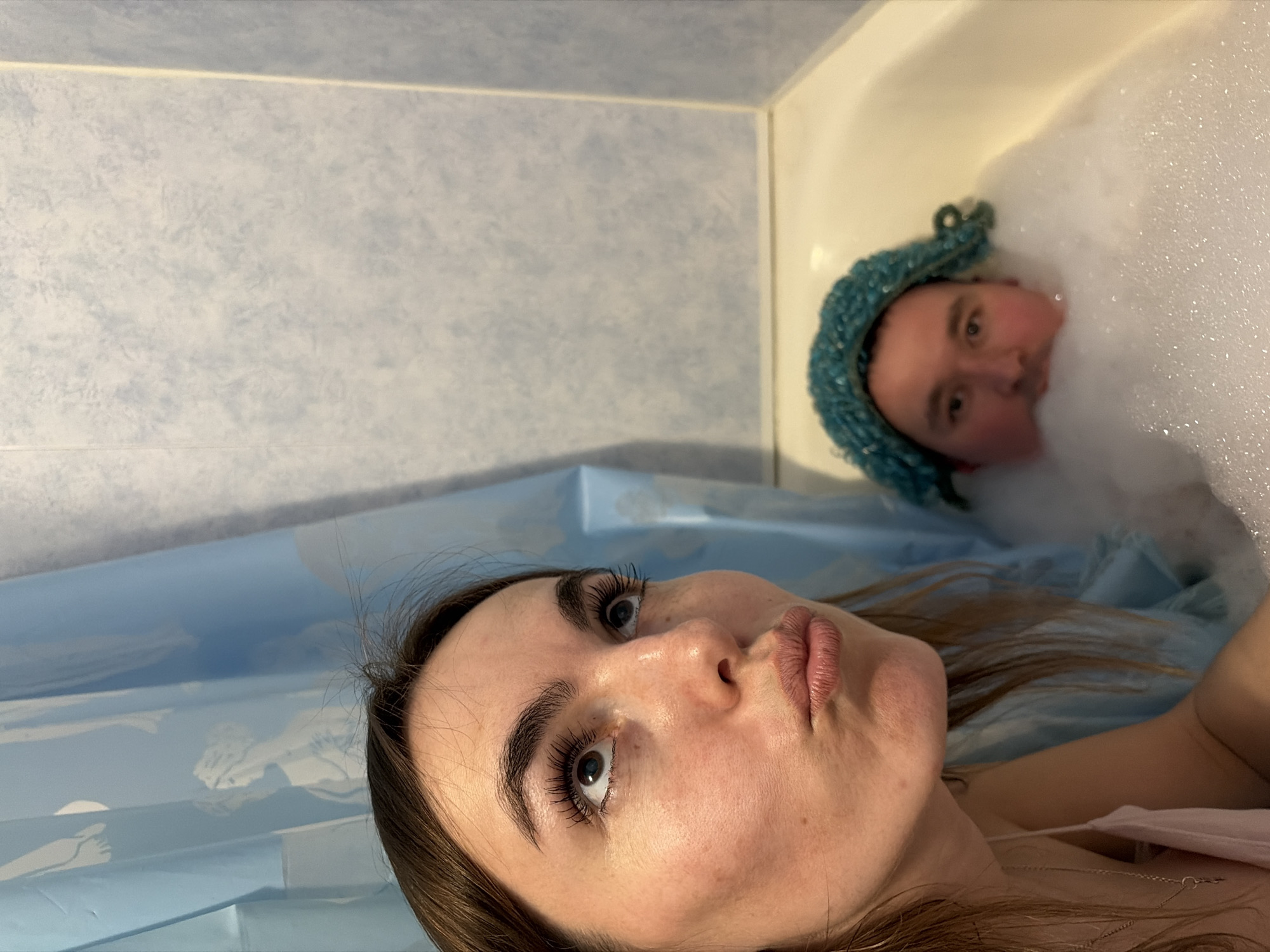 Two people are in a bathroom. One person, a brunette woman with long eyelashes, is visible from the chest up, lying down in front of a blue shower curtain. The other person is in a bubble bath, wearing a blue crocheted hat. The location of the photo is unknown, and nothing in the image indicates it was taken in Izhevsk, Russia.