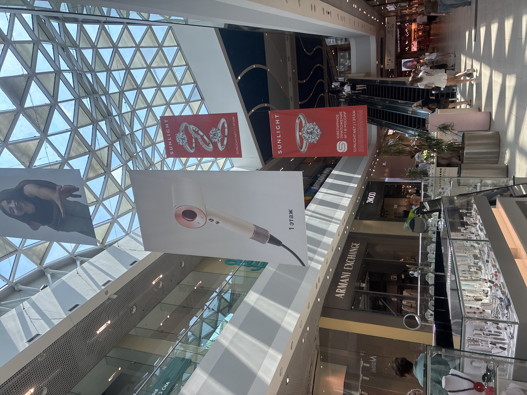 The image depicts the interior of the Galeria shopping mall in Sochi, Russia. Sunlight streams through a large, geometric glass roof, illuminating the multi-level atrium. Banners hang from the ceiling, advertising "SUNLIGHT" jewelry stores. One banner features an image of a Dyson hair dryer. Below, storefronts are visible, including "ARMANI EXCHANGE" and "2MOOD". People are seen walking, shopping, and using an escalator. The scene is a snapshot of a bustling commercial environment on a bright, sunny day.