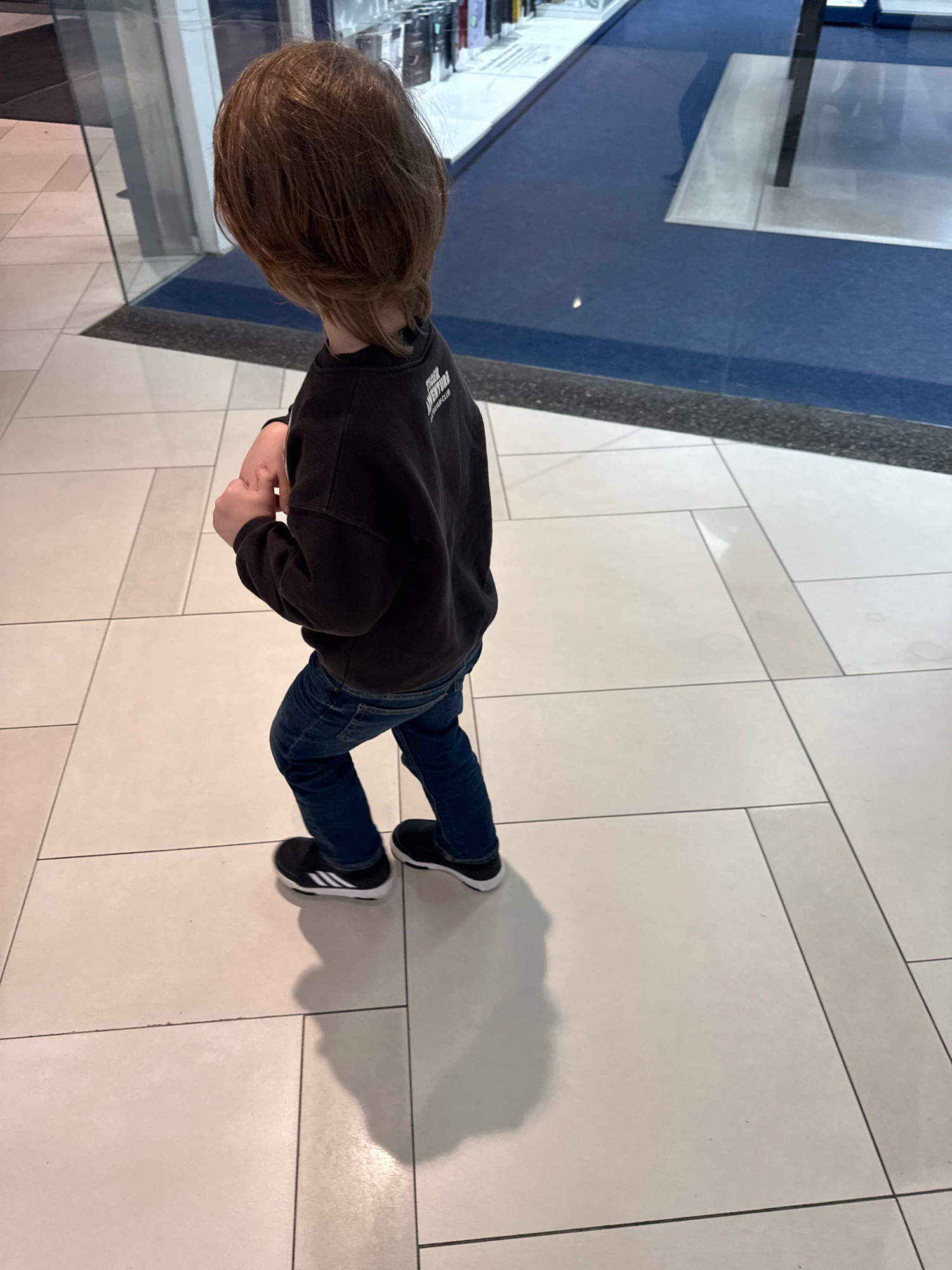 A child with brown hair, facing away from the camera, stands on a tiled floor. The child wears a dark long-sleeved shirt with text on the back and dark blue jeans. The child's feet are clad in black slip-on shoes with white stripes. The background features a reflective surface and a blue carpeted area. The scene is likely indoors, possibly in a commercial establishment or public space. The lighting suggests an indoor environment with artificial light sources. The floor is composed of large, light-colored square tiles with dark grout lines. A distinct shadow of the child is cast on the floor to the right. The specific location in Tallinn, Estonia cannot be determined from the visual information alone.