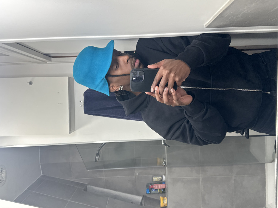 A person is seen in a bathroom mirror, taking a selfie with a smartphone. They are wearing a black zip-up hoodie and a bright blue hat. The bathroom features gray tiling, a shower head, and toiletries on a shelf. The mirror reflects part of the room, including a white wall and vent.