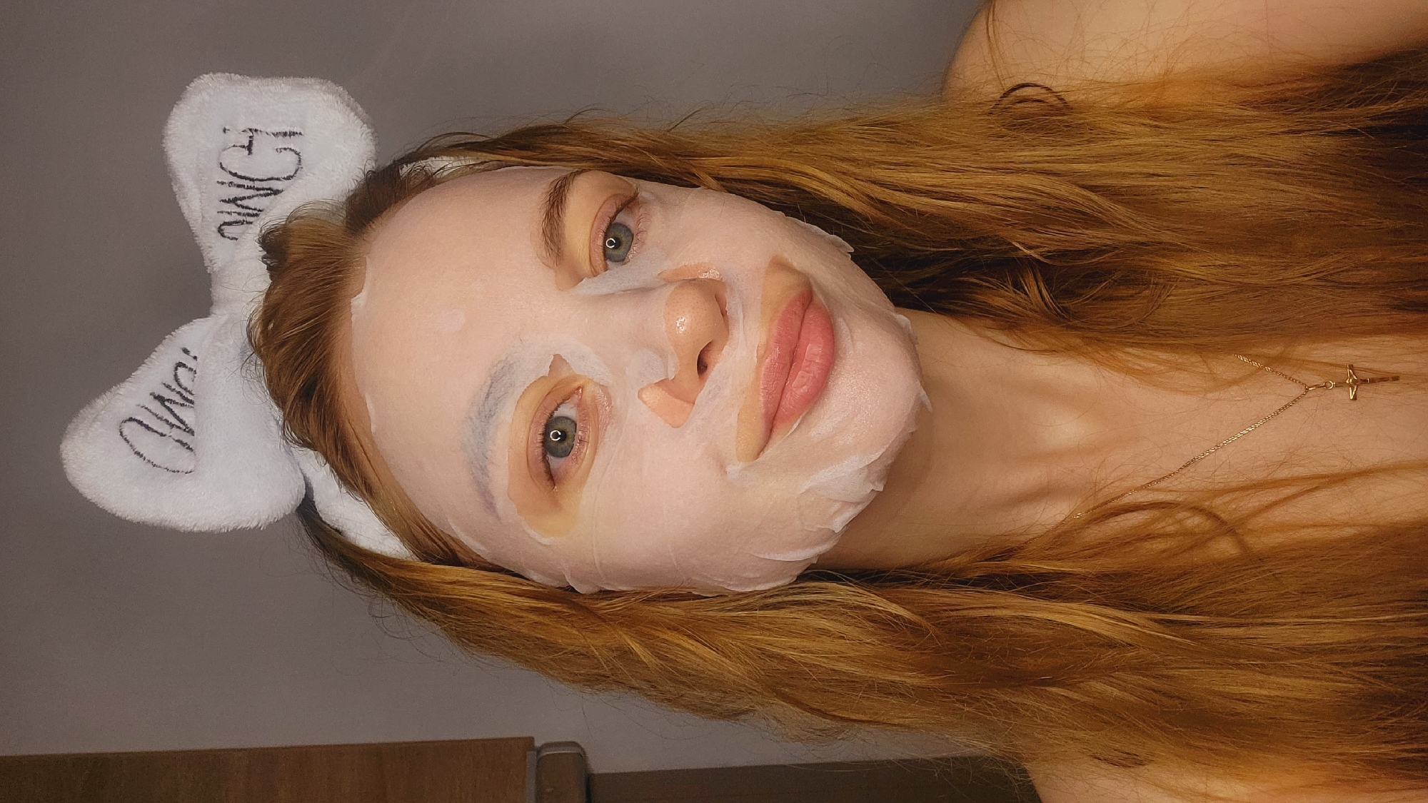 A person with red hair wearing a white sheet mask and a white headband with black lettering is lying down. The person is fair-skinned with light eyes, and a gold necklace is visible on their neck. The background appears to be a wall with a wooden cabinet partially visible. The location is not discernible as Chaikovsky, Russia.