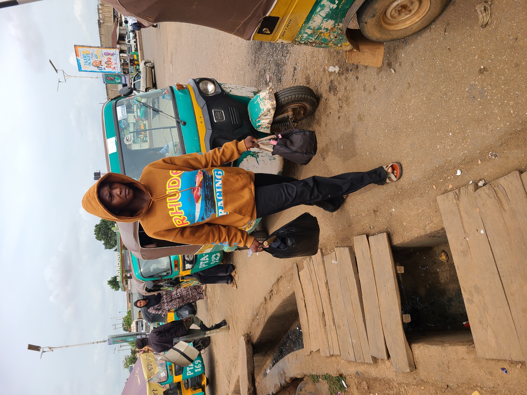 A man stands in front of a green and yellow vehicle, possibly a tuk-tuk, holding two bags. He wears a brown hoodie with a blue graphic and black pants with red sandals. The ground is dirt and pavement, with a covered drainage ditch on the right. Other tuk-tuks are parked in the background, and other pedestrians are present. There is a billboard in the background. Based on the tuk-tuks and general surroundings, the location is likely a city in an African country. The sign indicates this is near the MainLand.