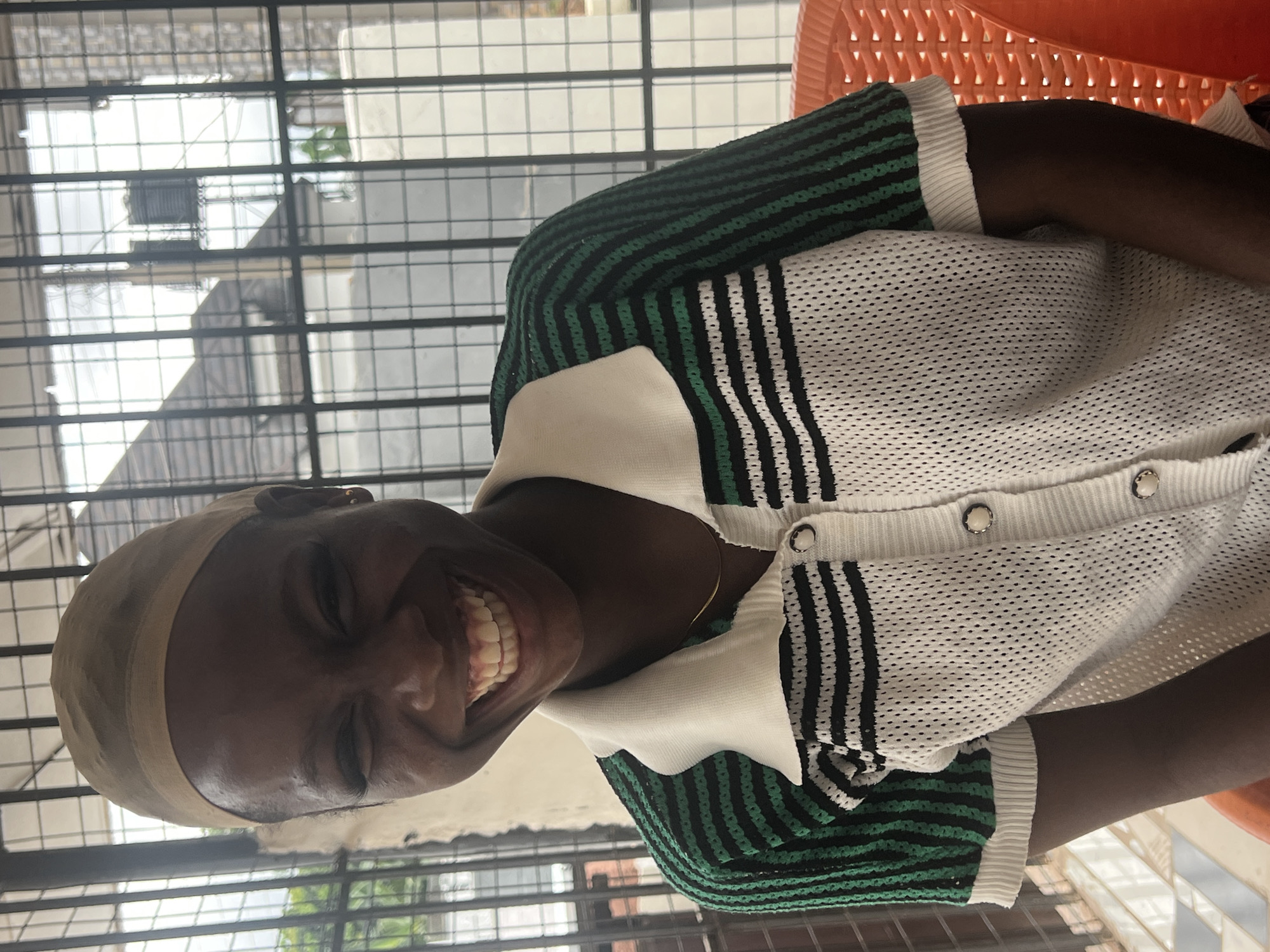 A medium shot photograph captures a Black woman with dark skin and a wide smile, revealing her teeth, wearing a beige wig cap. She has on a collared, short-sleeved cardigan with a white base and black and green stripes. The cardigan has buttons down the front. She is seated on an orange plastic chair in what appears to be an outdoor or semi-outdoor setting in Nkpor, Nigeria. Behind her, a barred window overlooks a residential area with a tiled roof, water tanks, and some greenery. The background also shows a concrete wall. The overall lighting suggests it is daytime.