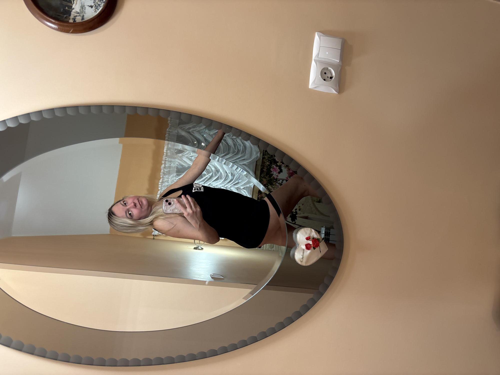 The image depicts a blonde female taking a mirror selfie in an interior residential space in Русский Вожой, Russia. She is shown from the waist up, wearing a black sleeveless top and dark lower garments, with a dark strap visible on her left thigh. Her right hand holds a smartphone with a clear case. The large oval mirror, framed with a scalloped grey border, reflects a light-colored wall, white draped curtains, and a floral pattern. Outside the mirror, the peach-beige wall displays a circular wooden-framed picture of a winter scene in the upper right, and a white electrical outlet with a single Schuko socket and two switches in the lower right. A white heart-shaped decoration adorned with red roses and the inscription 'С любовью' (With love) is attached to the bottom of the mirror.