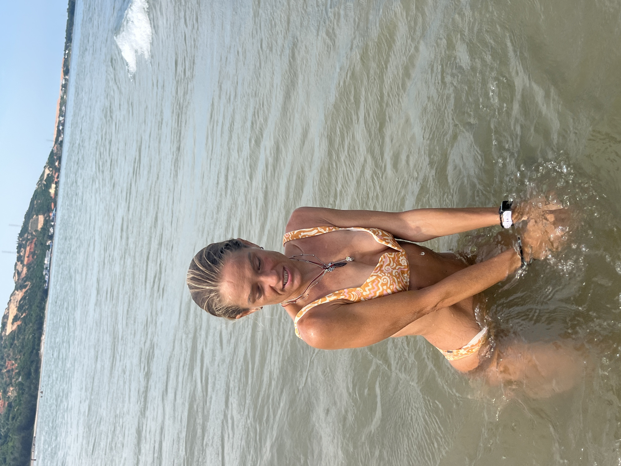 A woman with blonde hair pulled back is sitting in the ocean water, wearing a bikini with an orange and white patterned design. She is looking down with her mouth slightly open, and her hands are in the water in front of her. There is a necklace with a pendant around her neck, and a watch on her left wrist. The ocean water is a murky green color, with small waves and ripples. In the background, a distant shoreline with green hills and some buildings can be seen under a clear blue sky. This is a view of the ocean in Mũi Né, Vietnam.