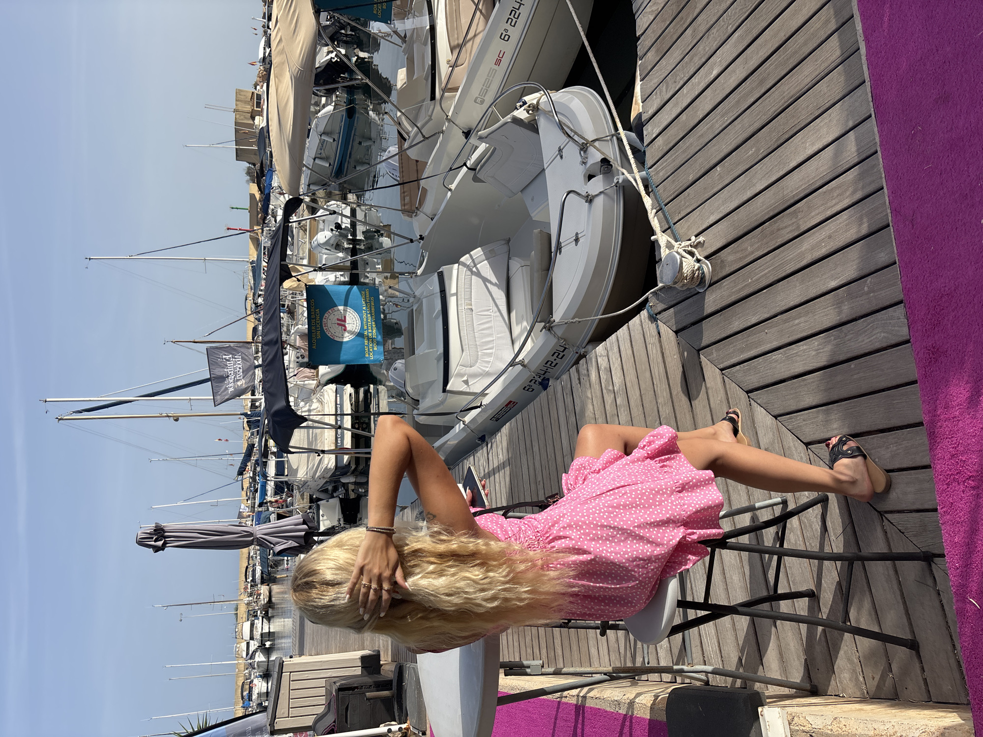 In Urbanización Cabo Roig, Spain, a blonde-haired woman with long, wavy hair sits on a stool at a round table on a wooden pier. She is wearing a pink polka-dot dress and black sandals. Her right hand is raised to her head, touching her hair, and she is holding a smartphone in her left hand. The pier is lined with various boats, including a white inflatable boat with a canopy in the foreground, and several sailboats and smaller motorboats docked behind it. In the background, there are more boats and buildings, and the sky is a clear blue. The scene appears to be a sunny day at a marina.