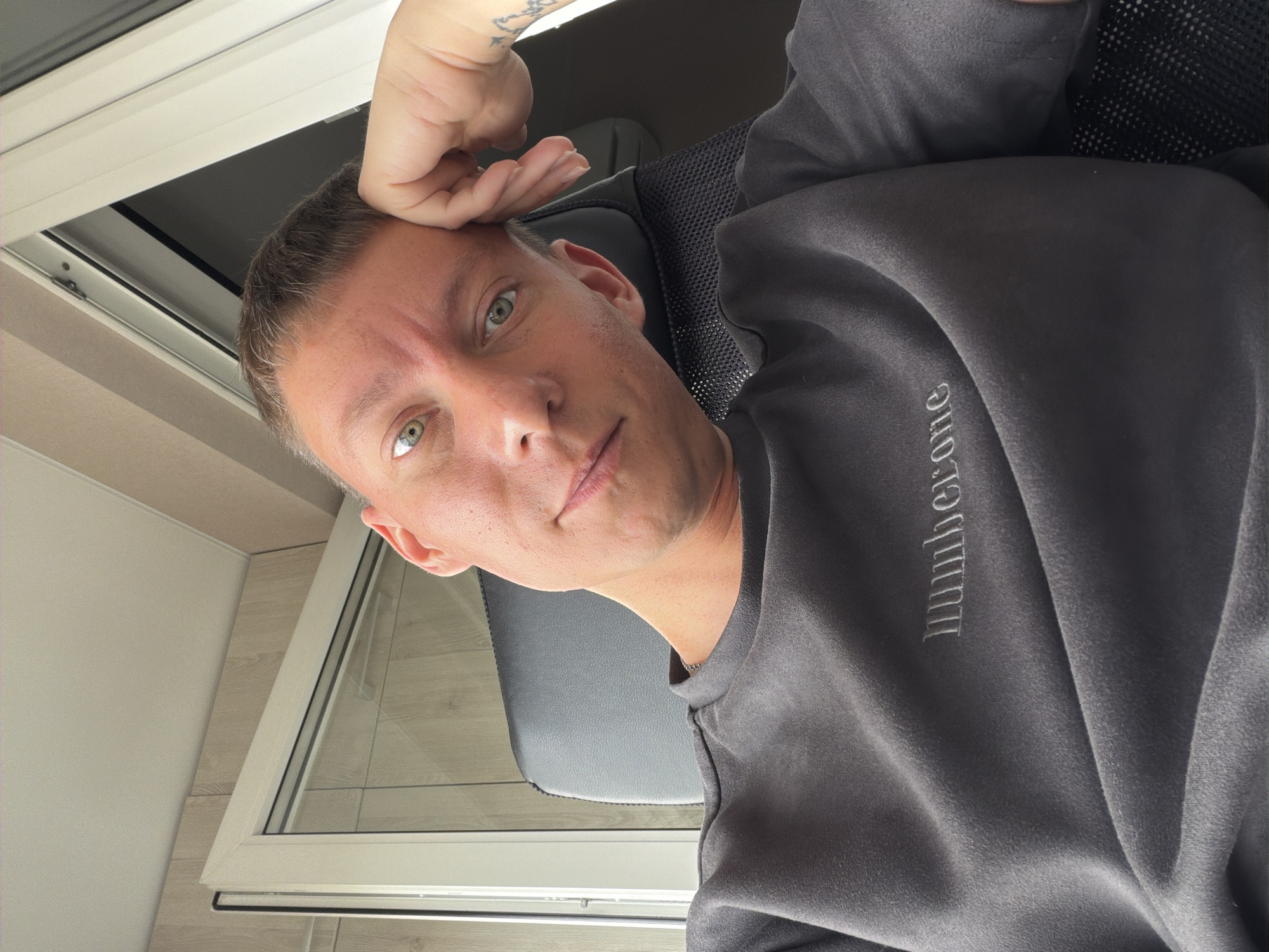 A selfie of a young adult male with light skin, short brown hair, and green eyes, taken indoors. He is wearing a dark gray sweatshirt with the word "Numberone" embroidered in silver thread on the left side of the chest. His left hand is resting on his forehead, and his head is tilted slightly to the right. He is seated in a dark gray office chair. Behind him, a white framed window with blinds is visible, and the wall behind the window appears to be covered in light-colored wood paneling. The lighting suggests it is daytime. No specific landmarks indicating Moscow, Russia, are present in the image.