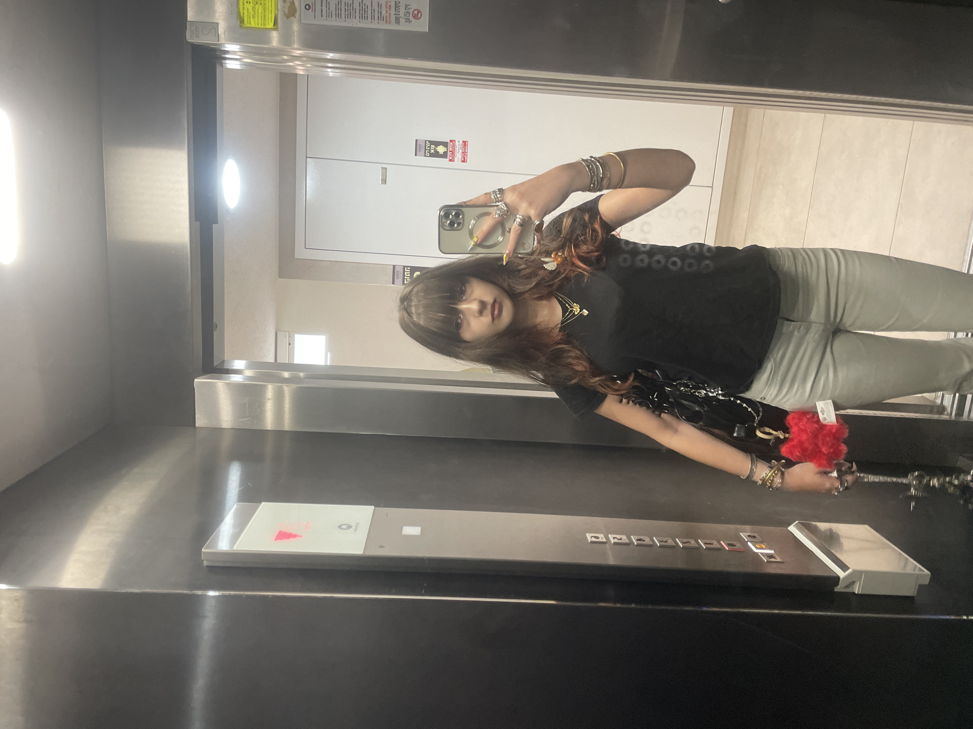 A young woman with bangs and reddish-brown hair is taking a mirror selfie in an elevator in Raanana, Israel. She is wearing a black t-shirt and light-colored pants. She has multiple rings on her fingers and bracelets on her wrists. She is holding a phone in her right hand, and a red fuzzy keychain is attached to a bag she is carrying. The elevator has a stainless steel panel with buttons and an illuminated display showing the number "3". The elevator doors are open, revealing a hallway with white cabinets and tiled flooring.