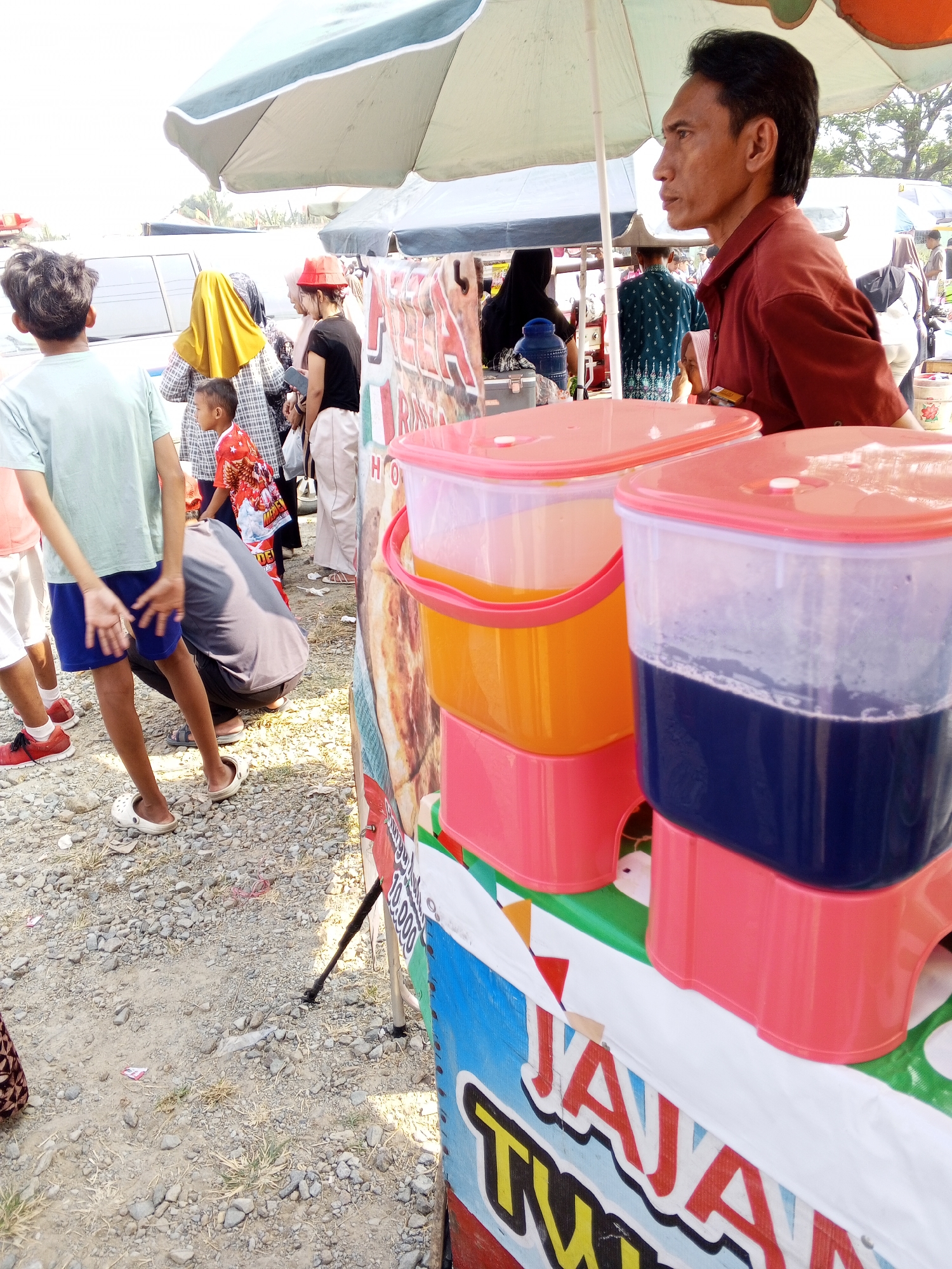 The image depicts an outdoor market scene in Ciruas, Indonesia. In the foreground, a vendor is seen from the side, a man with dark hair wearing a red shirt, standing behind a cart. On the cart are two large plastic drink dispensers with pink lids. The dispenser on the left contains orange liquid, and the dispenser on the right contains dark blue liquid. The cart itself is colorful, with blue, red, and white signage. In the mid-ground, several people are visible, including a young boy wearing a red shirt with a cartoon print, and a man in a grey shirt bending down. To the left, a young person wearing a light blue t-shirt and dark blue shorts is standing, facing away from the camera. In the background, more people and market stalls are visible under a large umbrella. The ground is covered with gravel and dirt.