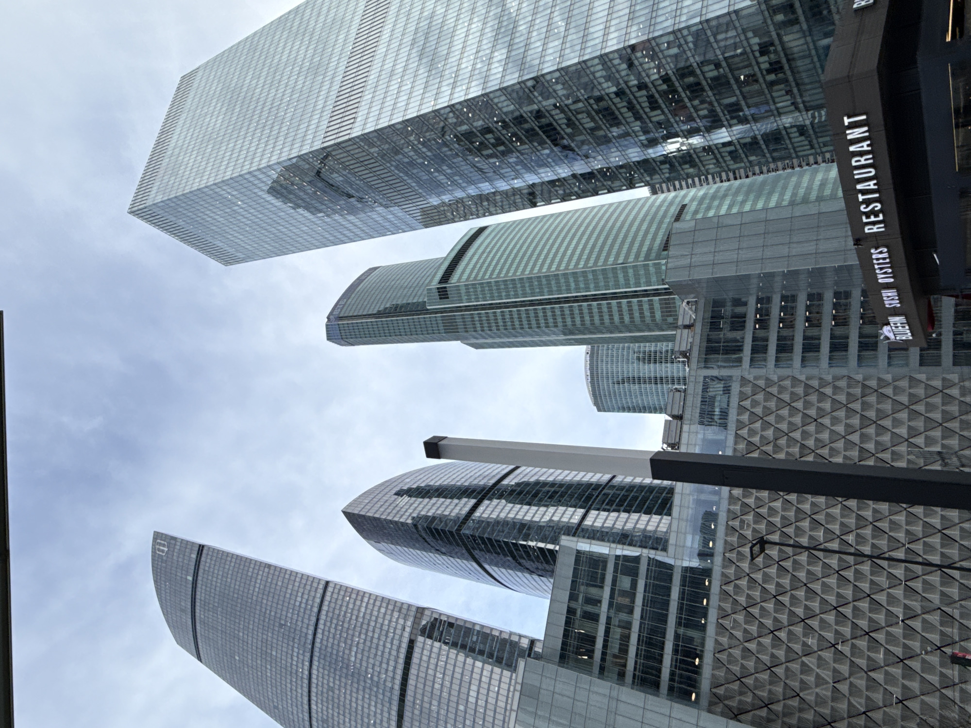The image depicts a low-angle view of several modern skyscrapers in a business district. The buildings are primarily constructed of glass and steel, with reflective facades. A sign for a restaurant called "BLUEFIN SUSHI OYSTERS RESTAURANT" is partially visible on the right side. The sky is overcast. The specific location in Khimki, Russia cannot be confirmed from the image.