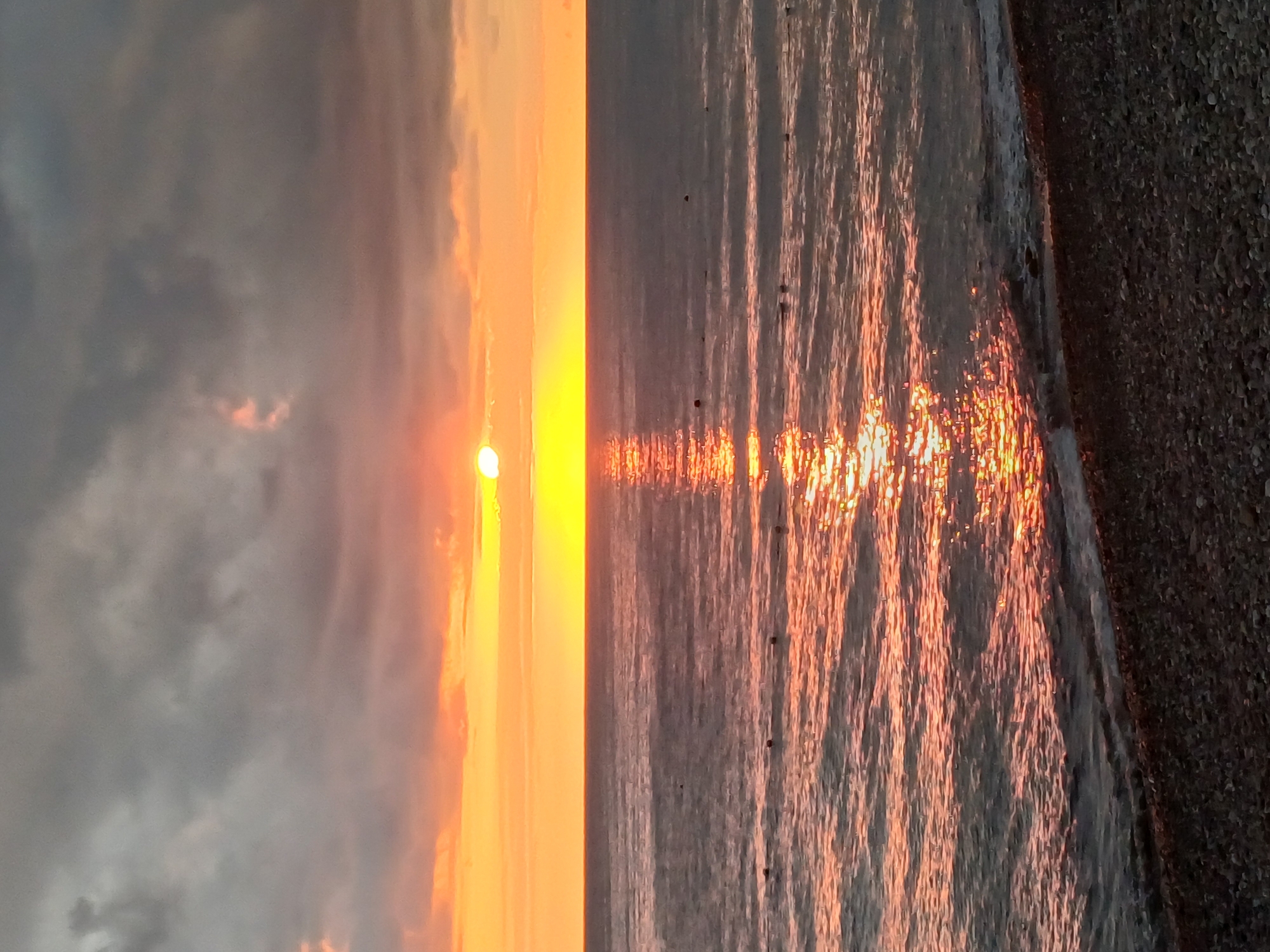 A vertical, eye-level, full shot captures a tranquil sunset over the Black Sea, likely near Sochi, Russia, given the typical seaside pebble beaches and the calm, relatively warm waters. The sun, a brilliant white orb, is partially obscured by dark, heavy clouds that dominate the upper portion of the frame, but it casts a warm, golden-orange glow across the sky and reflects vibrantly on the water. The sea is calm, with gentle waves lapping at the shore. A string of buoys, marked by small dark spheres, stretches across the middle distance of the water, possibly indicating a swimming zone or a demarcated area. The foreground is a pebble beach, dark and textured, with small, rounded stones. The overall atmosphere is serene and peaceful, with the dramatic sky and reflective water creating a visually stunning, yet calm, seascape.