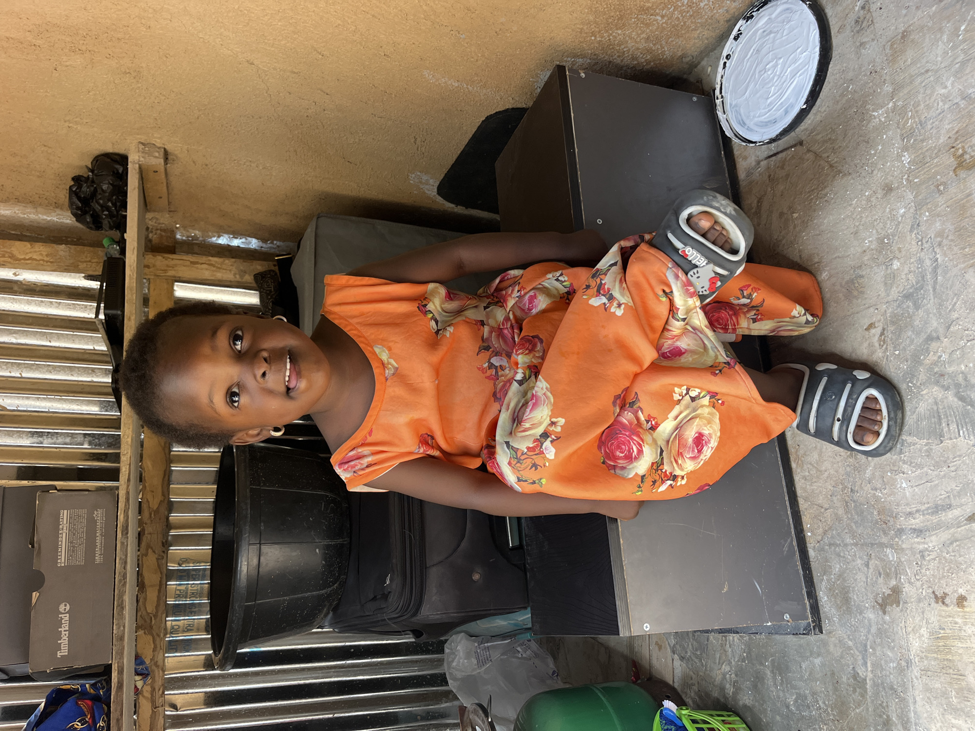 In an indoor, likely utilitarian setting, a young Black girl is seated. She is wearing a vibrant orange dress adorned with a floral pattern of pink and cream roses. Her hair is short and dark. She has on grey slip-on sandals with white trim. The girl is sitting on a dark grey, rectangular surface, possibly a storage container or a piece of furniture, which is placed on a grey, textured floor.
Behind her, stacked items are visible. To her left, a large black basin rests atop black luggage. Above this, a shelf holds a black box with "Timberland" branding and other writing. Further back, corrugated metal siding forms the wall. To her right, a textured, beige wall dominates the background, with a wooden shelf above it holding a black plastic bag and other indistinct items. On the floor to her right, a black, shallow container filled with a white substance, possibly paint or plaster, is visible. The overall environment appears somewhat cluttered and basic.