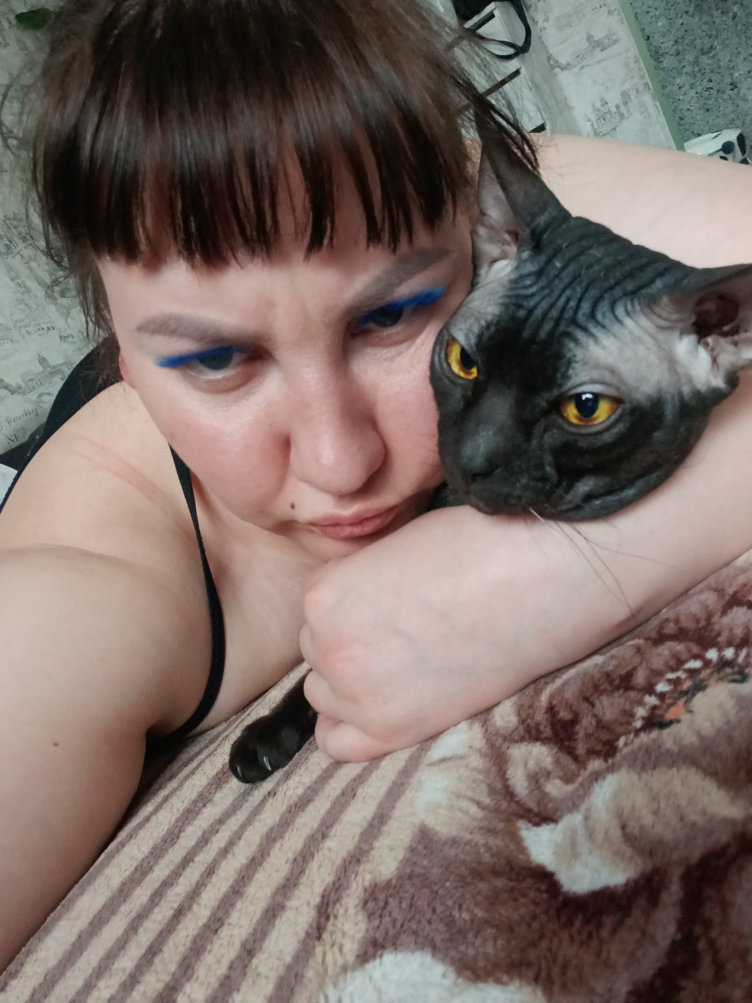 A woman with dark hair and blue eyeliner is lying on a striped blanket. A black and grey cat rests on her arm. The environment includes a patterned blanket and what appears to be wallpaper. The precise location in the city of Osa, Russia is not discernible from this image.