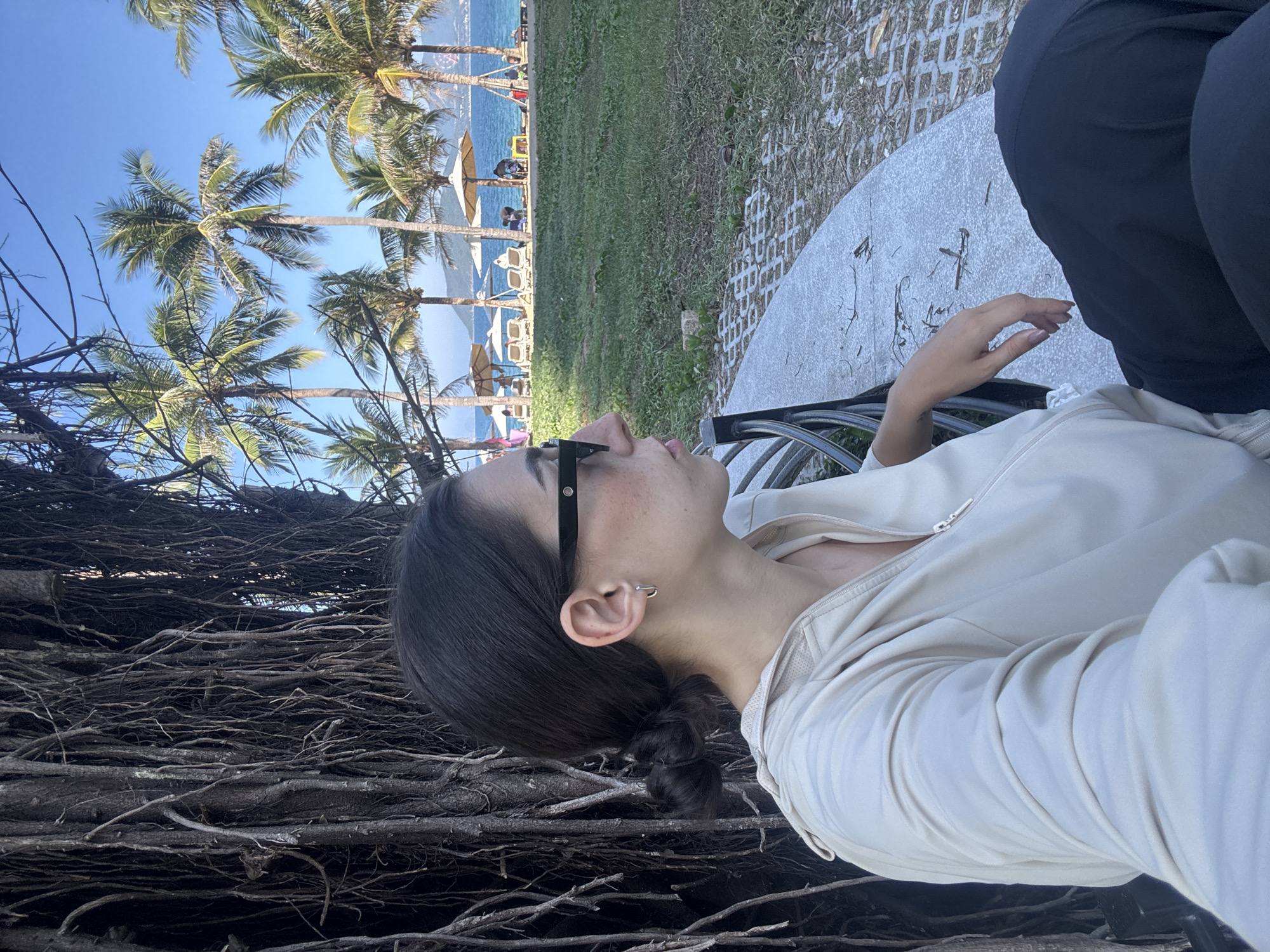 The image depicts an outdoor scene in Nha Trang, Vietnam, on a clear, sunny day. In the foreground, on the left, a person is seated on a curved grey concrete bench with a black metal frame, positioned in profile facing right. The individual has dark hair styled in a low bun, wears dark sunglasses, a light beige long-sleeved zipped top, and dark trousers. A small silver hoop earring is visible on the right ear. The right hand rests casually on the lap. To the immediate left of the person, a large tree trunk with numerous dark, hanging aerial roots forms a dense, textured background.
To the right of the bench, a strip of sparse green grass with patches of dry earth and a section of paved ground with a grid pattern is visible. Beyond this middle ground, a beach and ocean environment unfolds under a bright blue sky. Several tall palm trees with green fronds are prominent along the beach line. The light blue sea stretches into the distance, where a hazy landmass is discernible on the horizon. On the sandy beach, multiple light-colored beach umbrellas and sun loungers are arranged, with several indistinct figures of people present, some appearing to be relaxing or walking.