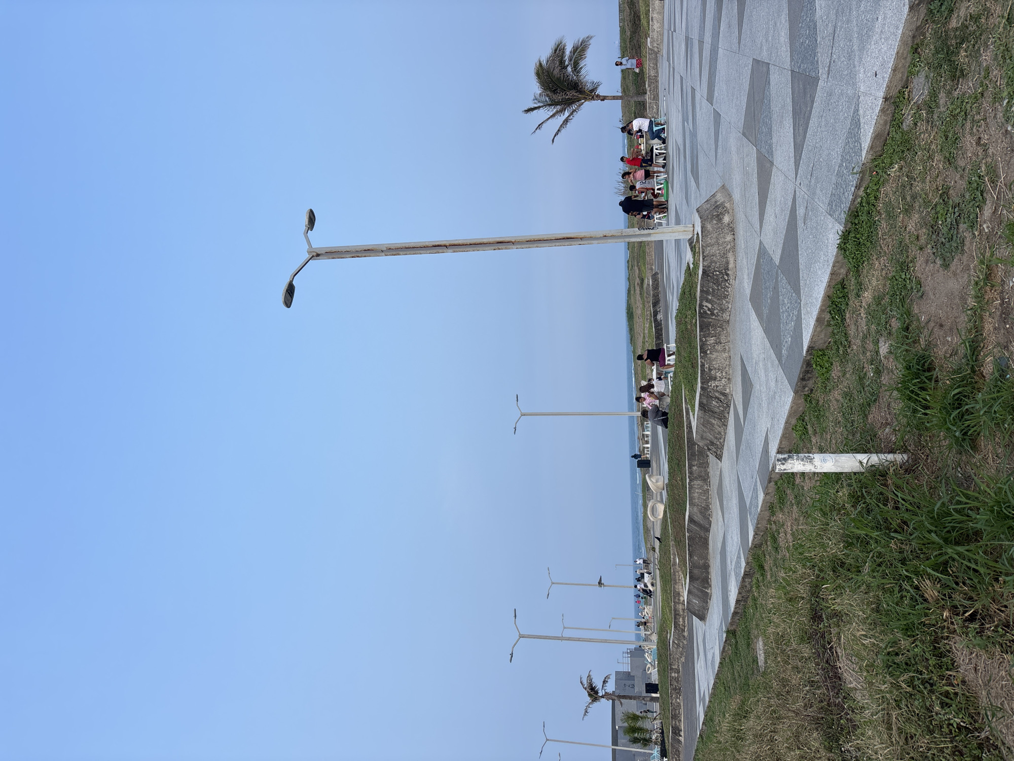 The image shows a sidewalk with a patterned gray and white surface, adjacent to a grassy area, running along the coastline in Veracruz, Mexico. The sky is a clear light blue. Numerous light poles line the sidewalk. People are seated in chairs near a palm tree on the sidewalk. In the distance, the ocean is visible.