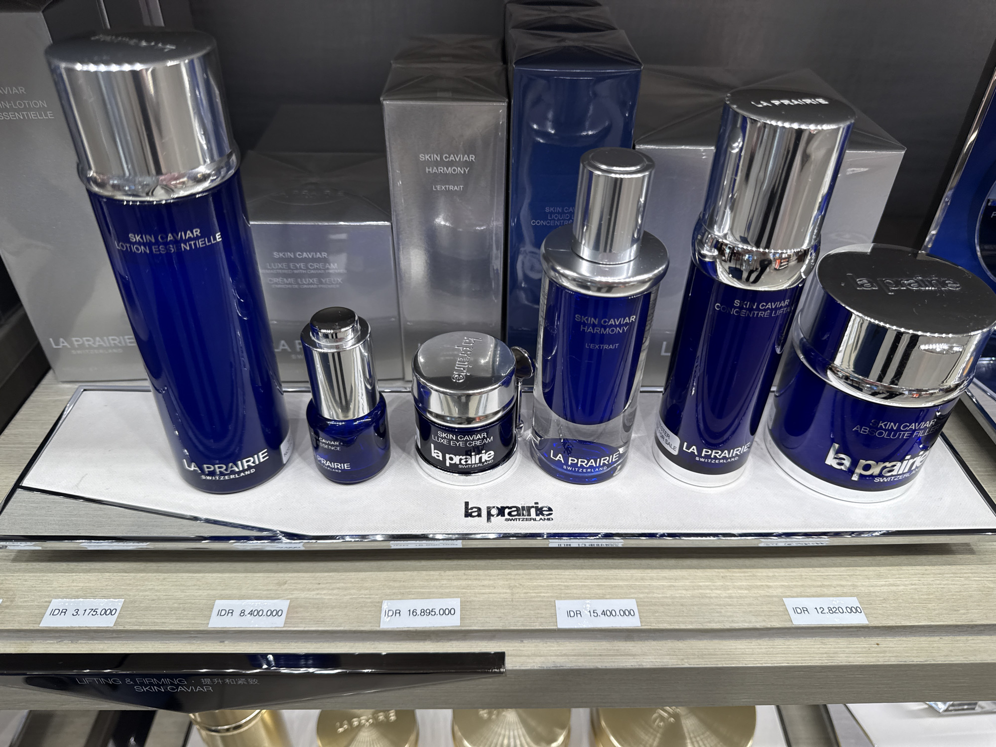 In a retail store in Kuta, Indonesia, a display of La Prairie skincare products is presented on a wooden shelf. The products, primarily in blue and silver packaging, are arranged in a staggered manner. Prominently featured are several bottles and jars of "Skin Caviar" products, including "Lotion Essentielle," "Luxe Eye Cream," "Concentré Lift," and "Absolute Filler." Small price tags in Indonesian Rupiah (IDR) are visible below the products, indicating prices such as 3,175,000, 8,400,000, 16,895,000, 15,400,000, and 12,820,000. The background shows more product packaging, suggesting a well-stocked display. The lighting is even, illuminating the products for presentation.
