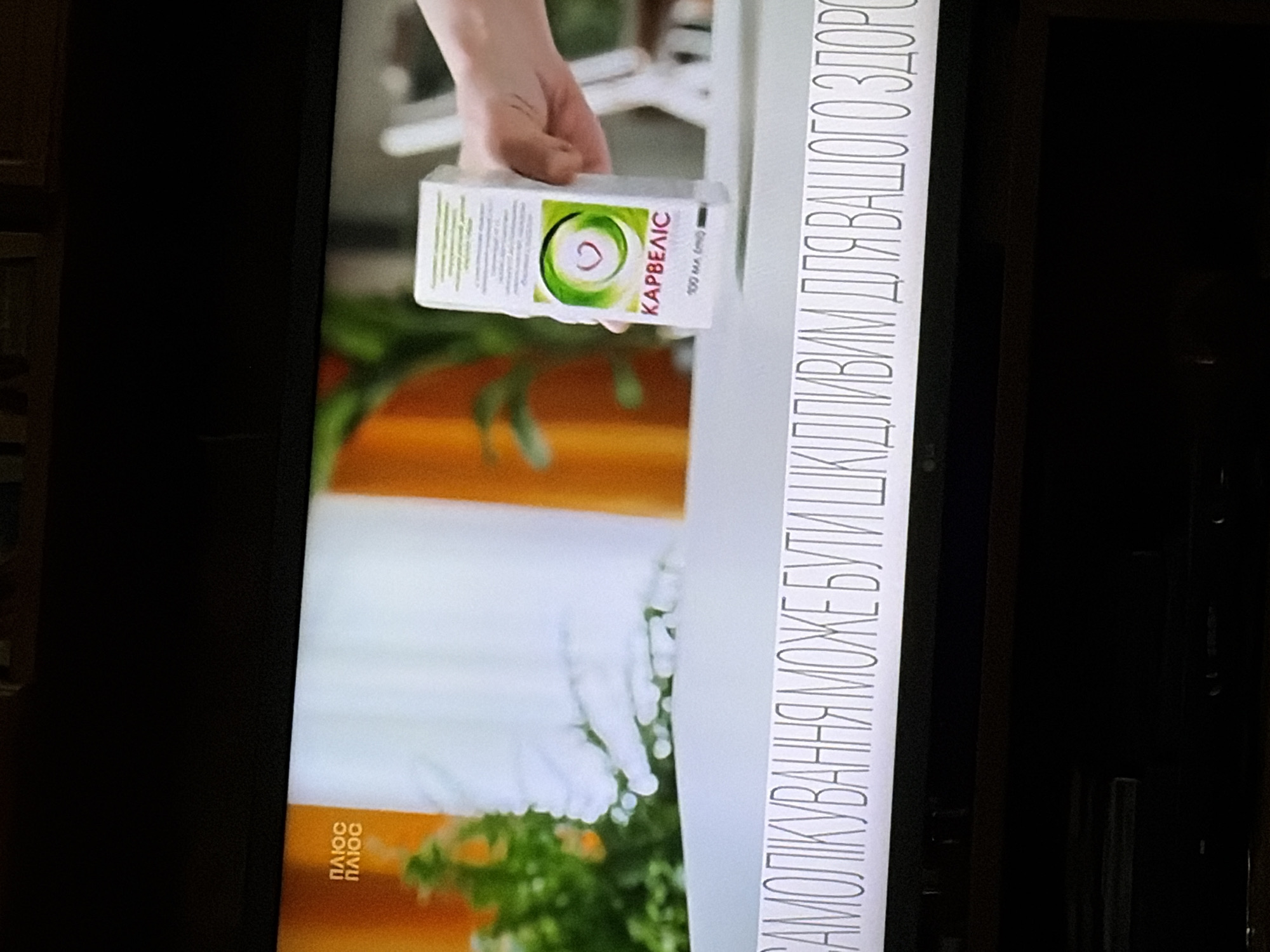 An overhead shot shows a TV screen displaying a segment of a program. On the left side of the screen, the words "ПЛЮС ПЛЮС" are visible in yellow text. A white box, likely a medication package, is held in a person's hand on the right side of the screen. The box has a green circular logo with a red heart in the center and the text "КАРВЕЛІС" below it. Below the box, a horizontal line of text in Ukrainian reads "САМОЛІКУВАННЯ МОЖЕ БУТИ ШКІДЛИВИМ ДЛЯ ВАШОГО ЗДОРОВ'Я," which translates to "Self-medication can be harmful to your health." The background of the screen features a blurry orange and green color scheme, suggesting a domestic or studio setting. The TV screen itself is positioned above a dark surface, which appears to be a piece of furniture.