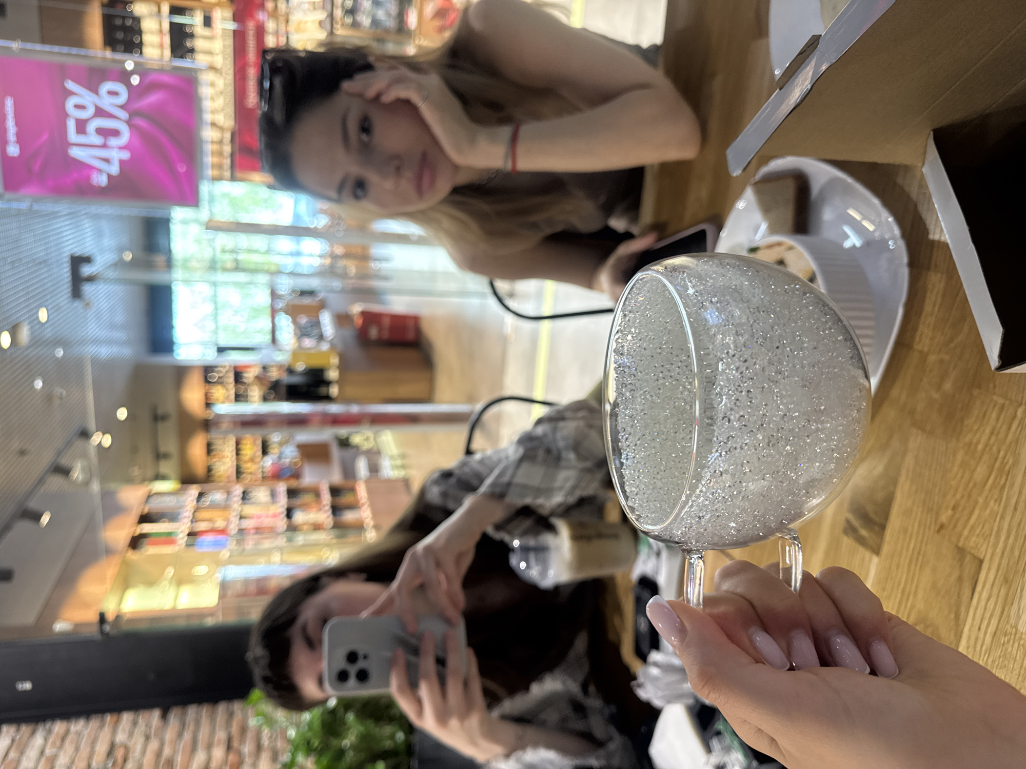 The image shows two women at a table in what appears to be a cafe or restaurant. One woman is posing for a photograph taken by the other. A large glass, seemingly filled with small, reflective objects, is held in the foreground by the person taking the picture. There is food on a plate nearby, along with a box. A sign with "45%" is visible in the background. The location is identified as the city of Moscow, Russia.