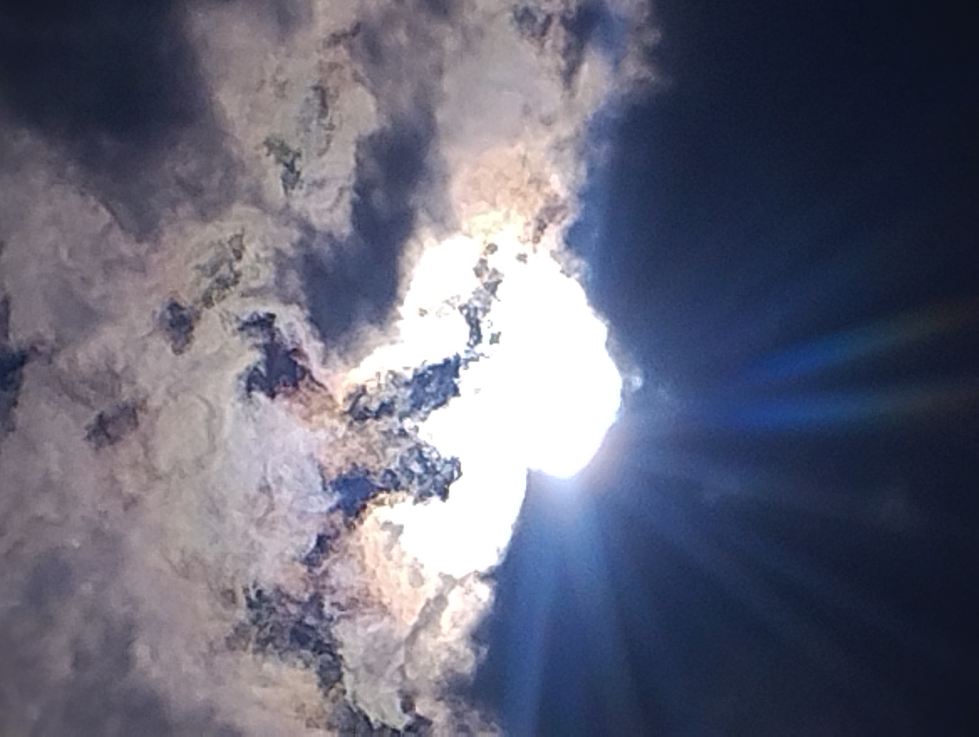 A bright sunburst pierces through dark clouds. The sun is a blinding white orb at the center of the image, with rays of light emanating outwards. The clouds are a mix of dark grays and whites, with some pink and yellow hues around the sun. The sky is a deep blue. The image appears to be a photograph taken from a low angle, looking up at the sky.