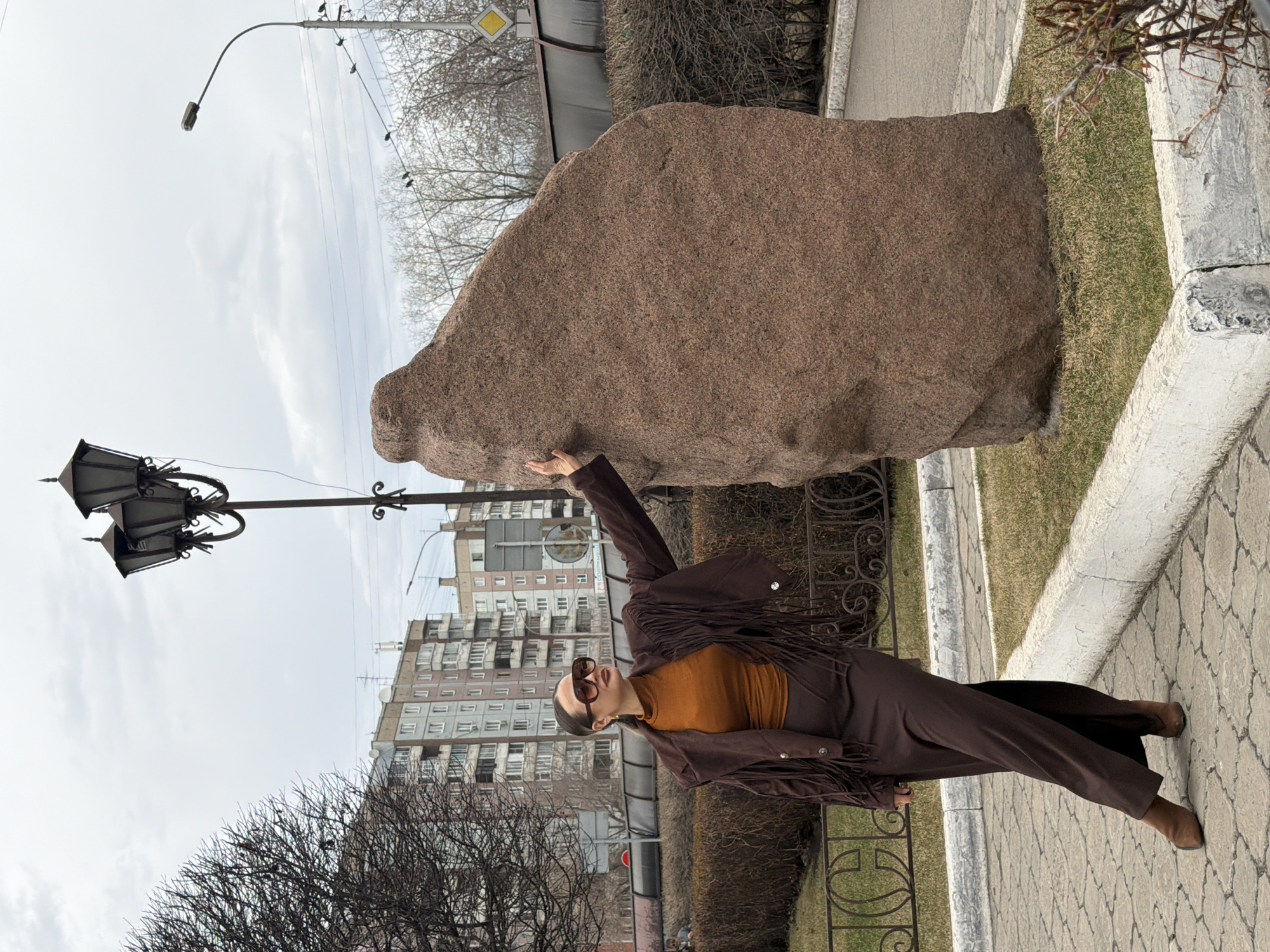 In an outdoor scene in Krasnoyarsk, Russia, a woman stands posing next to a large, reddish-brown rock with a rough texture. The image appears to be taken on an overcast day, likely in late winter or early spring, as indicated by the bare branches of trees and bushes. The woman is dressed in a brown jacket with prominent fringe details, an orange long-sleeved top, brown pants, and brown boots. She wears sunglasses and has her dark hair pulled back. Her left hand rests on the large rock as she faces slightly to the left.
The setting includes a paved walkway with a patterned surface. Behind the woman, a decorative, multi-headed street lamp with ornate metalwork stands tall. In the background, several multi-story residential buildings are visible, characterized by multiple windows. Further into the distance on the left, a simpler, modern street light on a curved pole is present. A black metal fence or railing separates the paved area from some of the background elements. To the right of the rock, a patch of light green grass is visible, bordered by a stone or concrete curb. A yellow diamond-shaped road sign can be seen in the upper right portion of the background. The sky is uniformly cloudy and bright. There is no visible text in the image.
