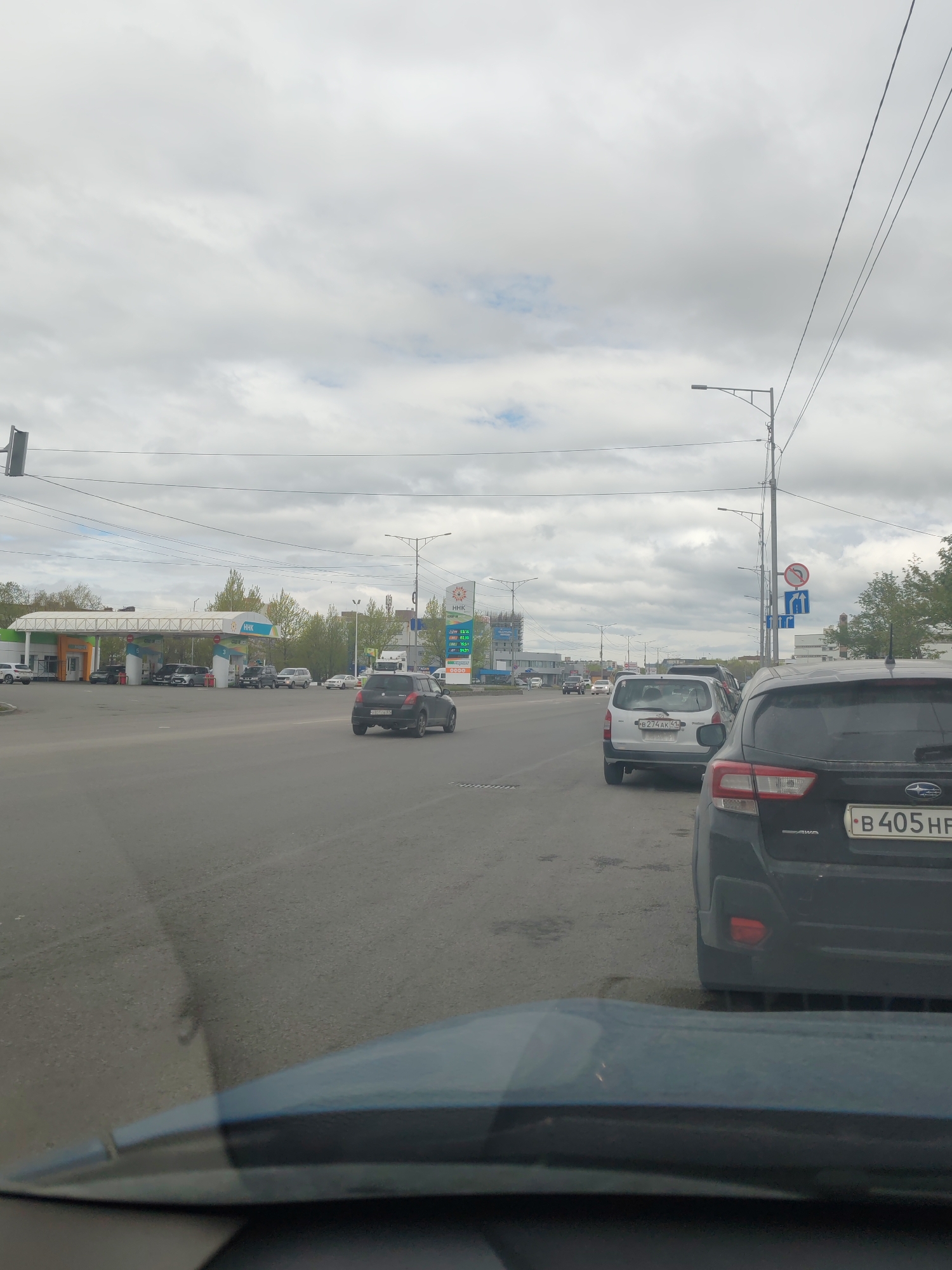 The image shows a view from inside a vehicle on a cloudy day, looking out at a street scene. In the foreground, the hood and windshield of the vehicle are visible. Further down the road, there is a black Subaru SUV with a license plate that reads "B 405 HF". To the left of the Subaru is a white hatchback, possibly a Suzuki, with a license plate that reads "B 274 AK 41". Beyond these vehicles, a gas station with the "HHK" logo is visible, with several cars fueling up. Traffic lights and street signs are also present. The environment appears to be an urban or suburban setting with trees and buildings in the background. The location is identified as Petropavlovsk-Kamchatsky, Russia.