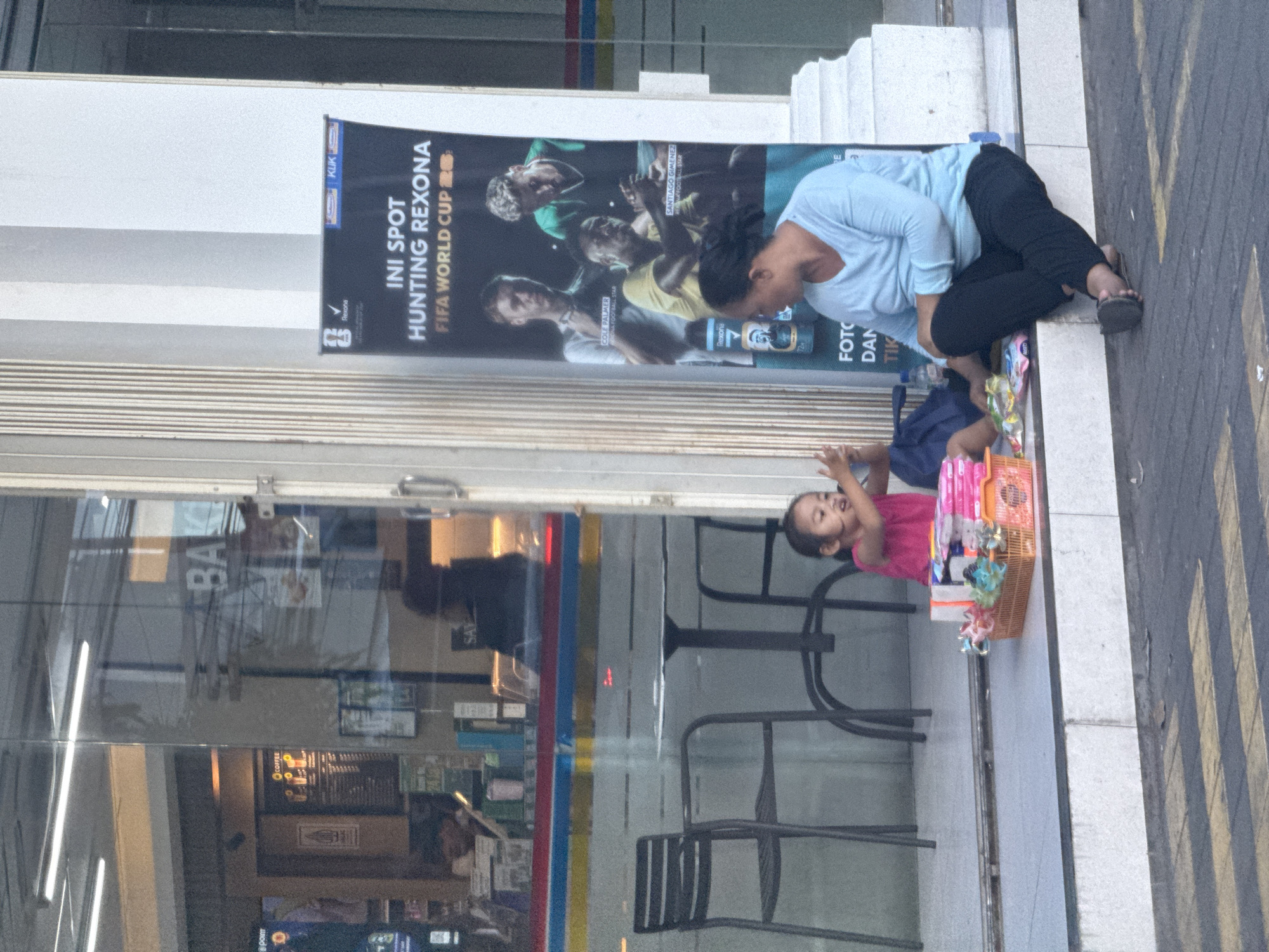 The image captures a candid street scene in Kuta, Indonesia, showing a woman and a young child interacting on a sidewalk next to a glass storefront. The woman, wearing a light blue long-sleeved top and black pants, crouches low, looking down towards the child or items on the ground. She wears dark sandals. Beside her, a young child in a bright pink top sits on the paved ground, looking up with a joyful expression and slightly raised arms. A woven basket filled with colorful small items, possibly toys or trinkets for sale, sits between the child and the woman, suggesting a street vending scenario.
The setting appears to be a busy commercial area, indicated by the large glass facade reflecting interior lights and furniture, including tables and chairs, possibly from a cafe or convenience store. The sidewalk features yellow markings, common in urban environments. Above the woman and child, a large black banner or poster is prominently displayed on the building.
Visible text on the banner includes "INI SPOT HUNTING REXONA FIFA WORLD CUP 22," featuring images of football stars like "COLE PALMER FOOTBALL STAR" and "SANTIAGO GIMENEZ REXONA FOOTBALL STAR," along with the brand "Rexona" and slogans like "FOTO DAN TIK" and "72H." Other partially visible text includes "KLIK," "COFFEE," "BAK," "SAY," and "Point," likely from signs within the reflected interior. The lighting suggests bright daylight. The overall mood conveyed is one of everyday life on the streets of Kuta, possibly a family engaging in street commerce.