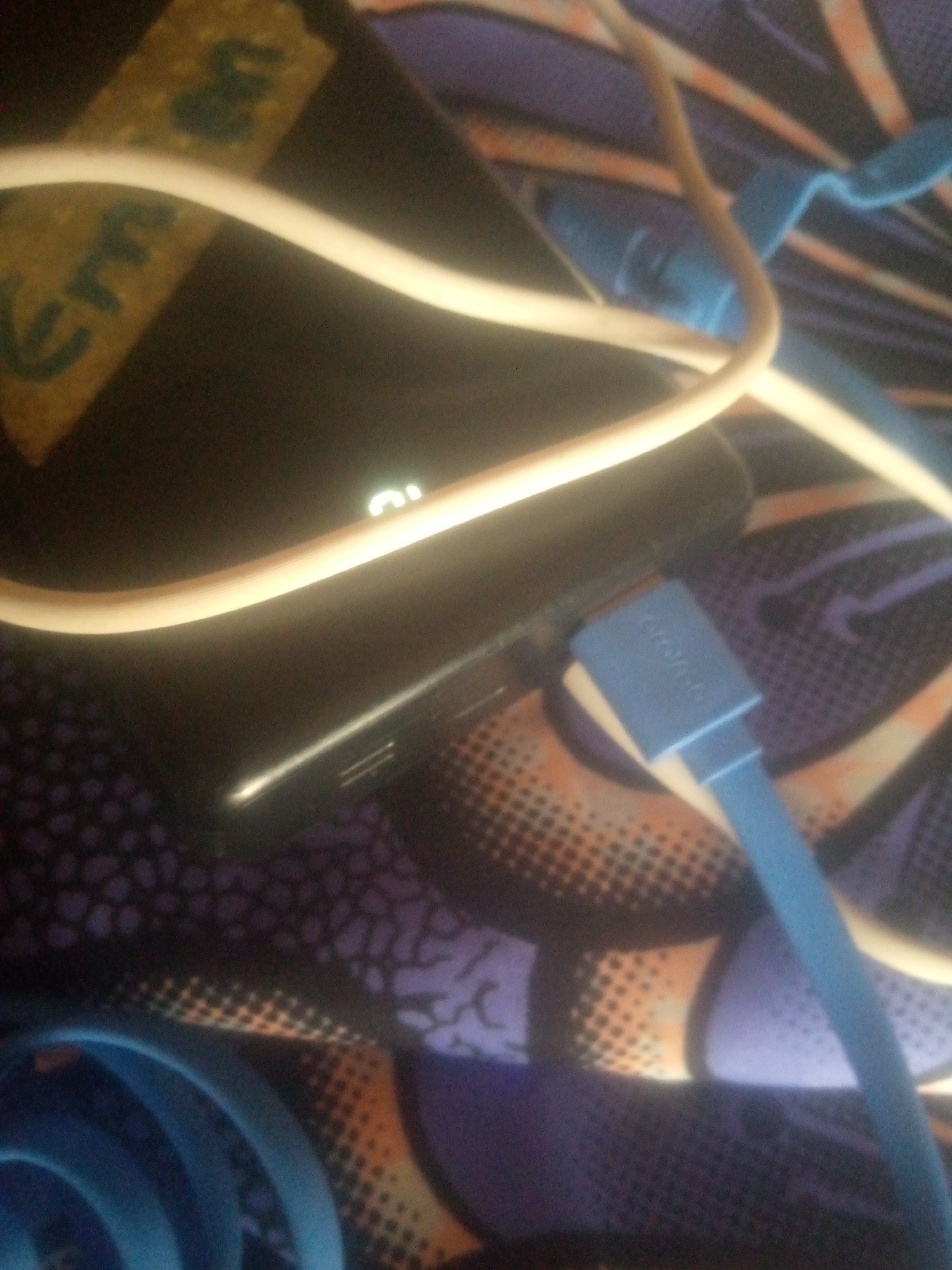 The image shows a close-up of electronic devices and cables on a patterned surface. A dark-colored rectangular object, likely a mobile phone or portable charger, is positioned diagonally. A white cable is draped across the object, and a blue cable with a USB connector is plugged into it. The blue connector has faint text that appears to say "Anker". Part of the patterned surface shows a design with purple, orange, and black elements, resembling a fabric or mat. In the upper left corner, a yellowed piece of tape with dark scribbled text is visible on the dark object. The lighting suggests an indoor setting, possibly with artificial light, as there is a bright highlight on the dark object and the cables. There are no people or clear indications of the time of day or weather. The context provided of Jauro Yinu, Nigeria, does not offer specific visual cues within the image itself.
